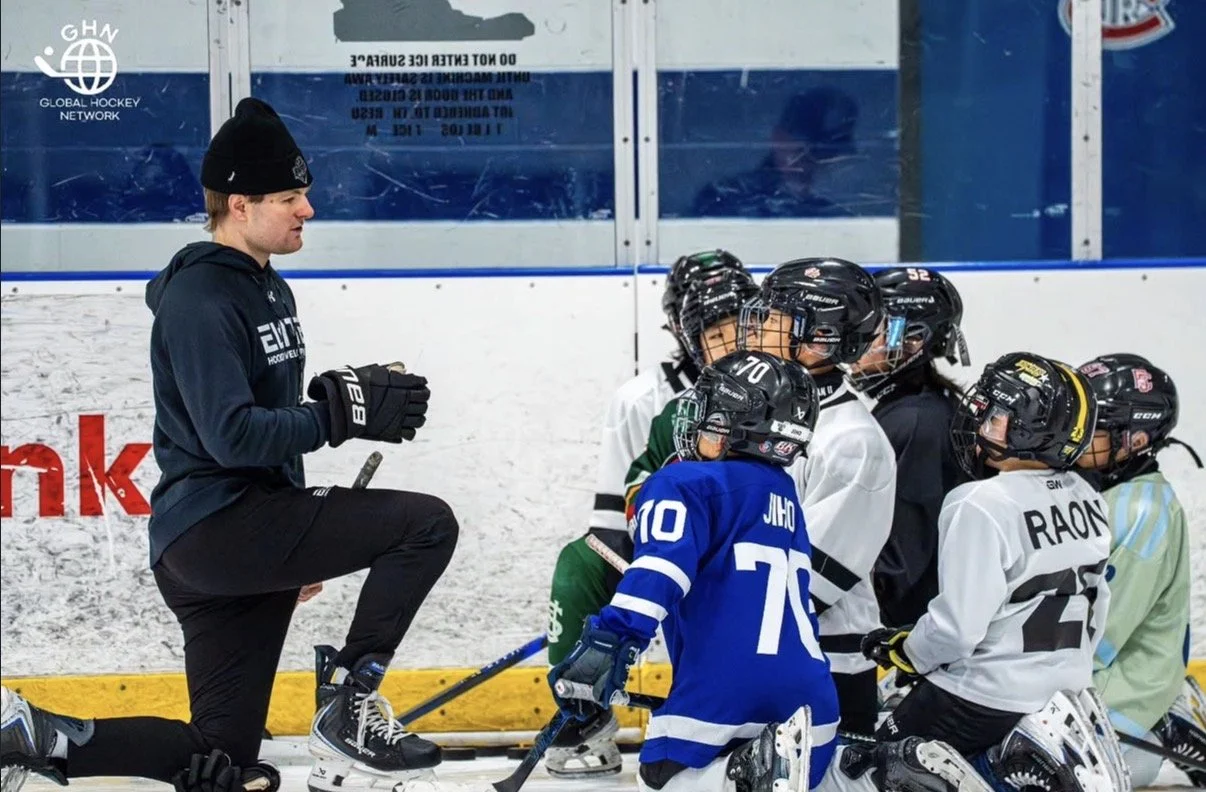 A hockey coach kneels on one knee on the ice rink, holding a hockey stick, addressing a group of young hockey players in full gear and helmets as they listen attentively during a practice session.