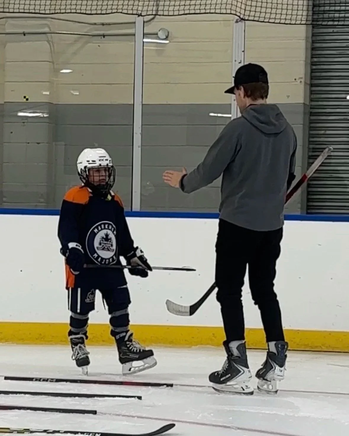 A young boy in hockey gear standing on an ice rink, listening to an adult man in casual clothes teaching or coaching him during hockey practice.