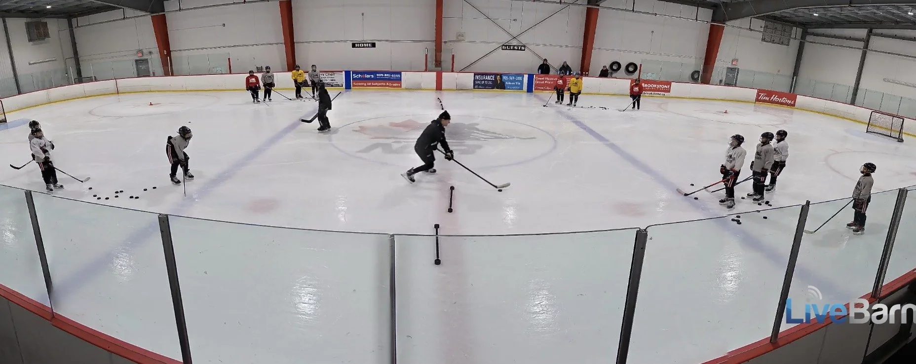 Children and coaches on an ice hockey rink during practice, with players in white and black, and coaches in darker clothing, practicing passing and skating.