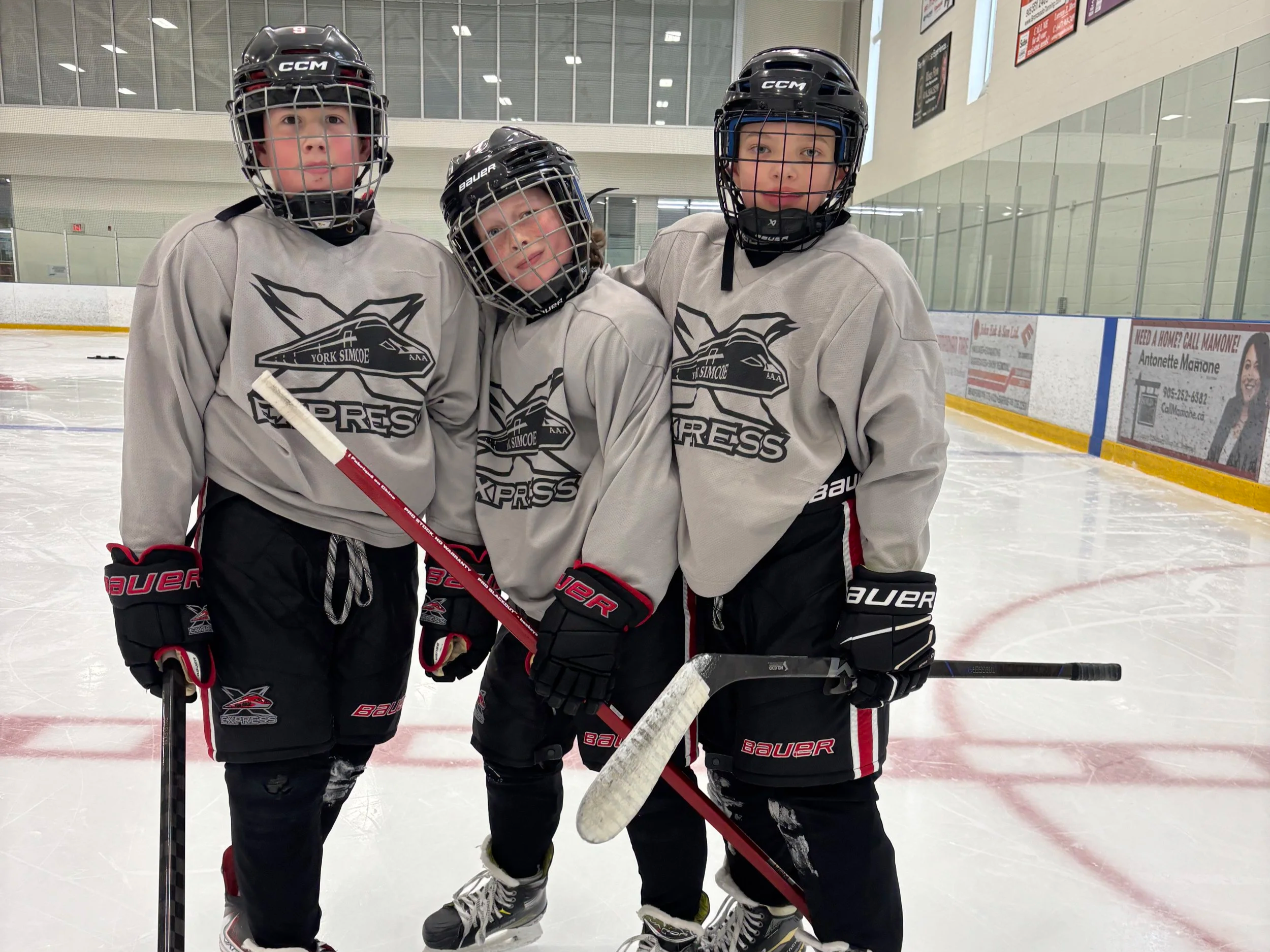Three young boys in hockey gear standing on an ice rink with arms around each other, wearing matching jerseys and black hockey helmets.