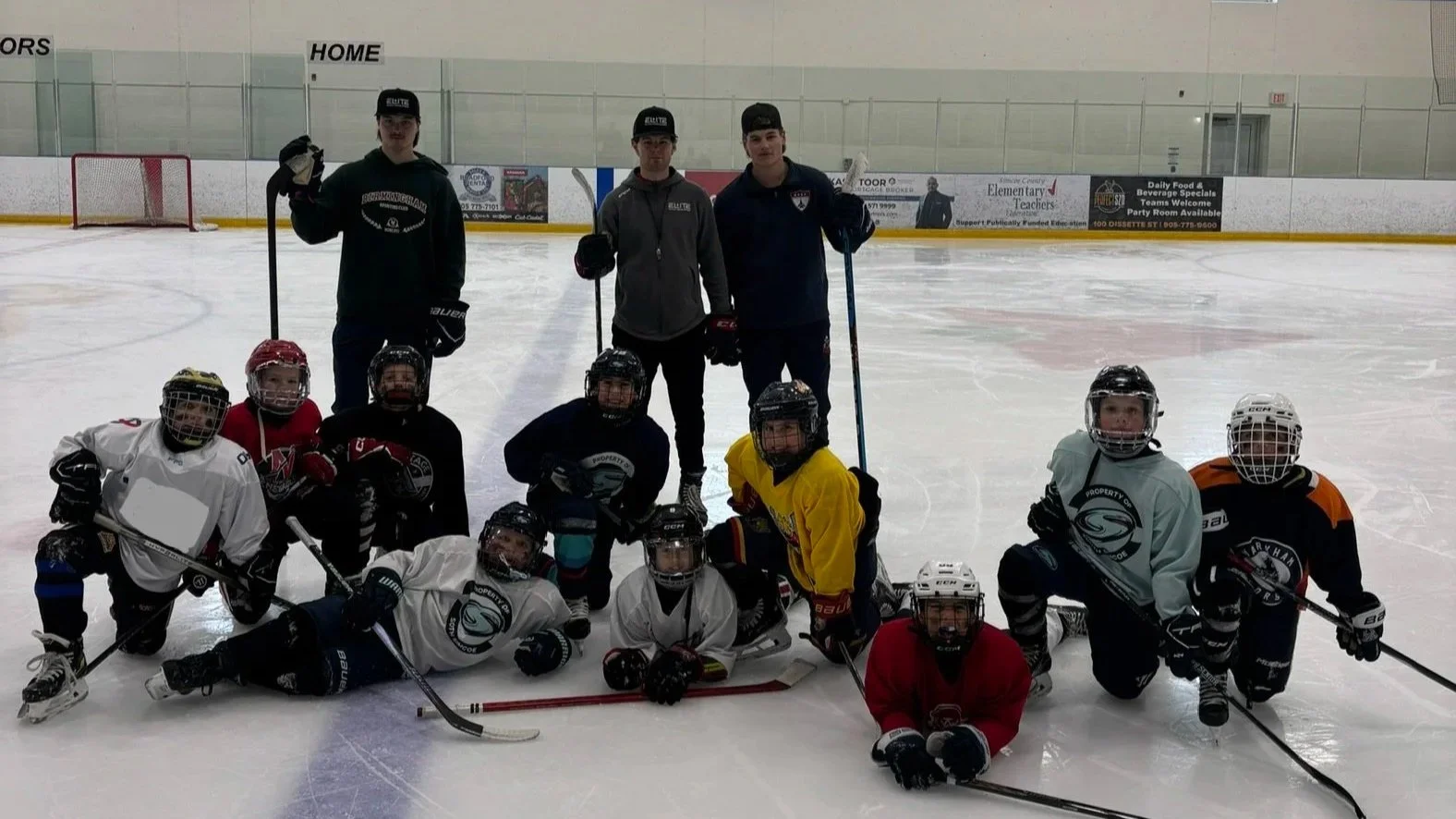 Group of children and coaches in an ice hockey rink, some children in hockey gear kneeling and lying on ice, coaches standing behind them, all holding hockey sticks.