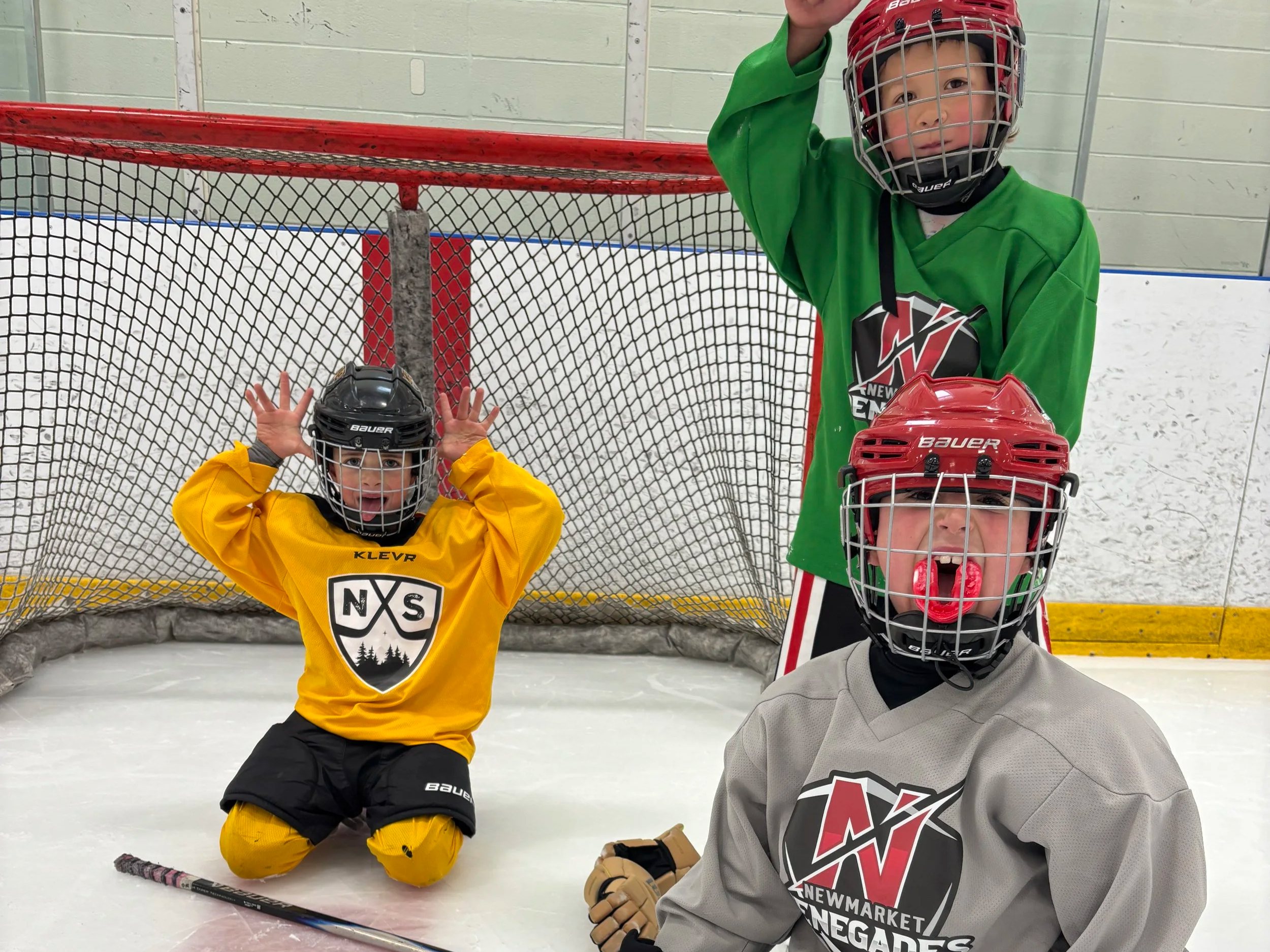 Three children in hockey gear on an ice rink, standing in front of the hockey net. One child in yellow hoodie is kneeling with hands on helmet, one child in gray jersey is sitting on the ice, and one child in green jersey is standing with a helmet and gloves. All are wearing helmets with face cages.