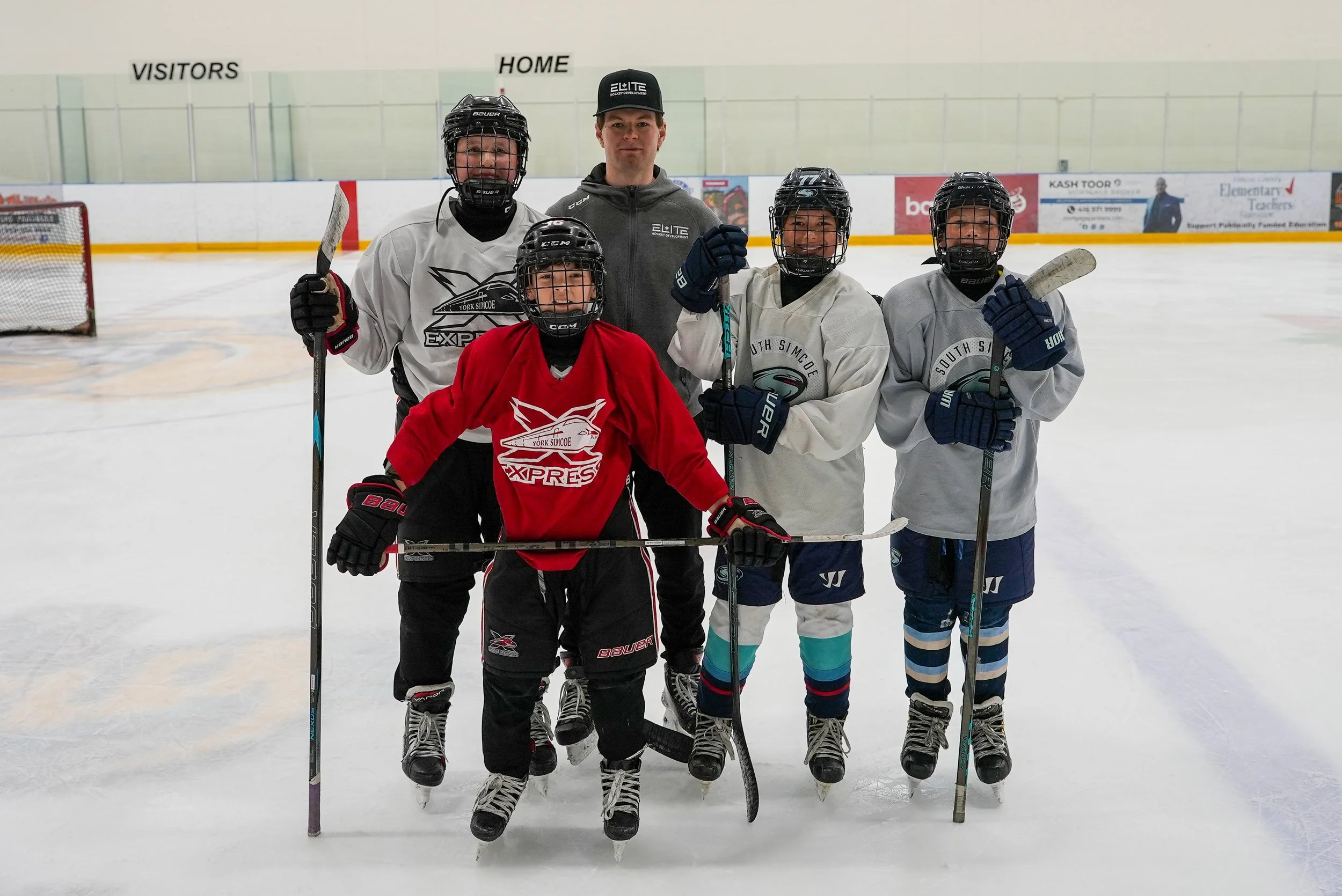 A group of five young hockey players and their coach on an ice rink, posing for a photo. The players are wearing helmets, gloves, and hockey gear. The coach is in the center, wearing a black cap and hoodie. The background shows the rink's boards and a sign indicating the visiting and home team areas.