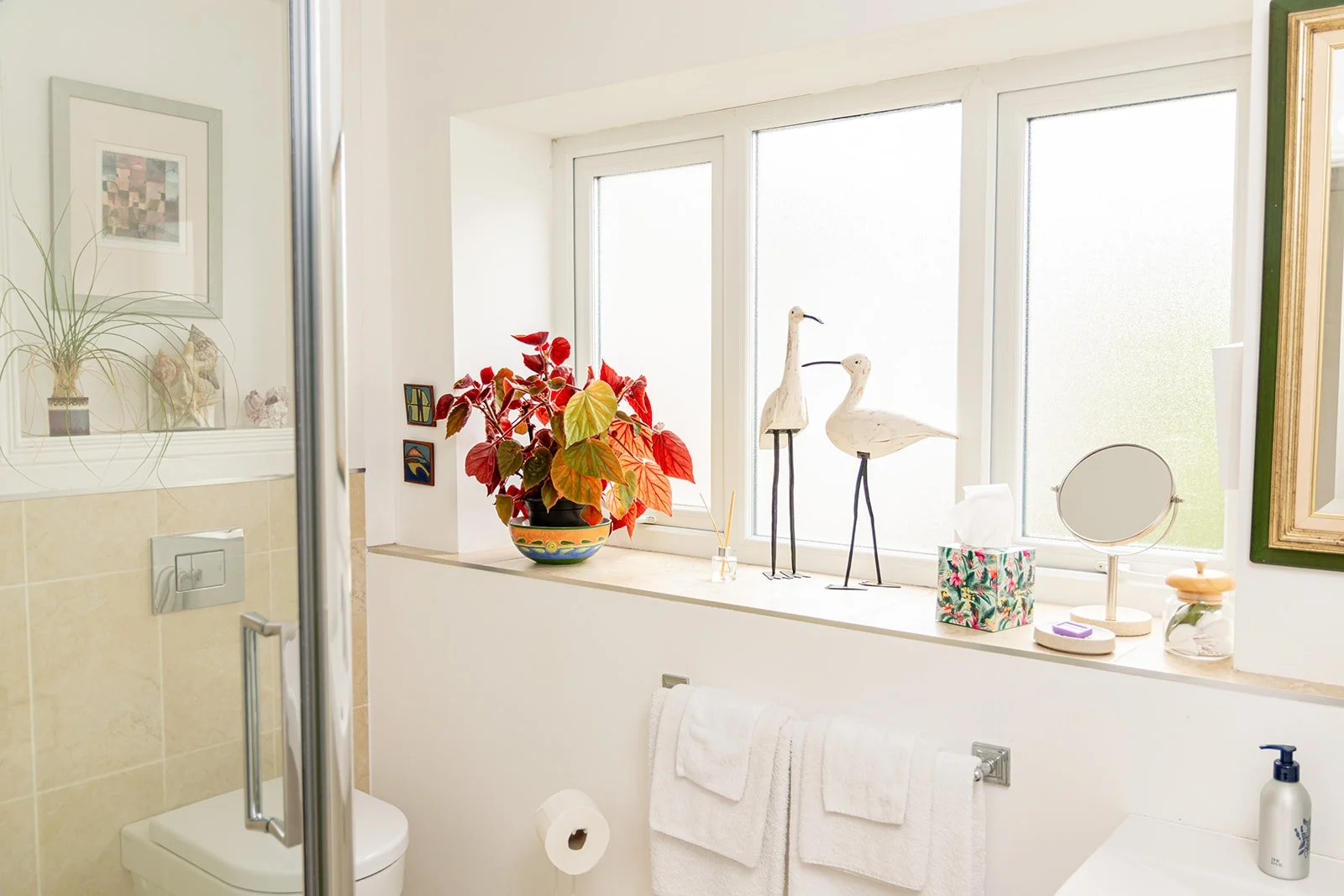 Bathroom window shelf with houseplant, decorative bird sculptures, tissue box, mirror, and various toiletries.