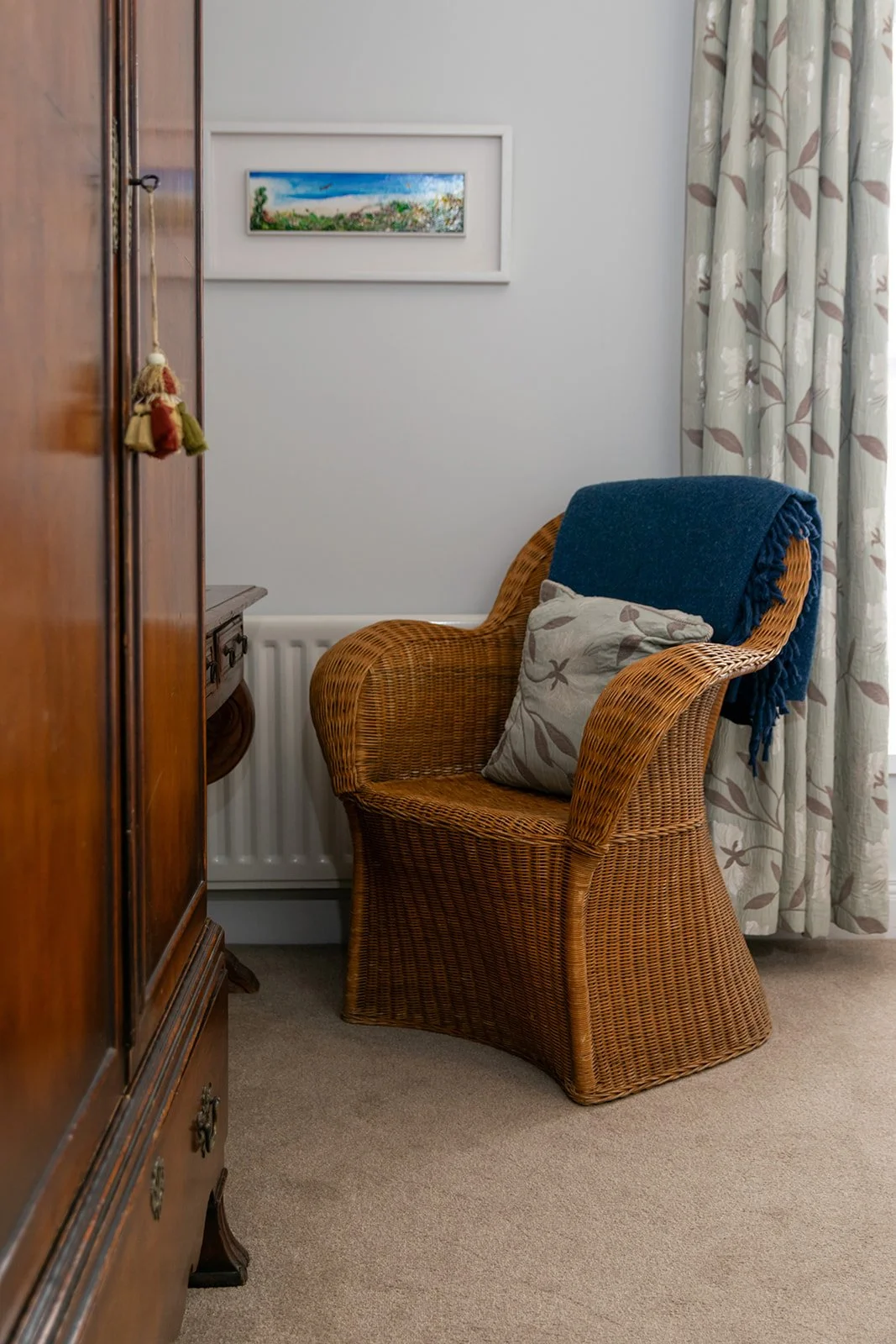 A cozy corner with a wicker armchair draped with a blue blanket, a cushion, and a view of a curtain with a floral pattern, a white radiator, a framed landscape painting, and a wooden cabinet.