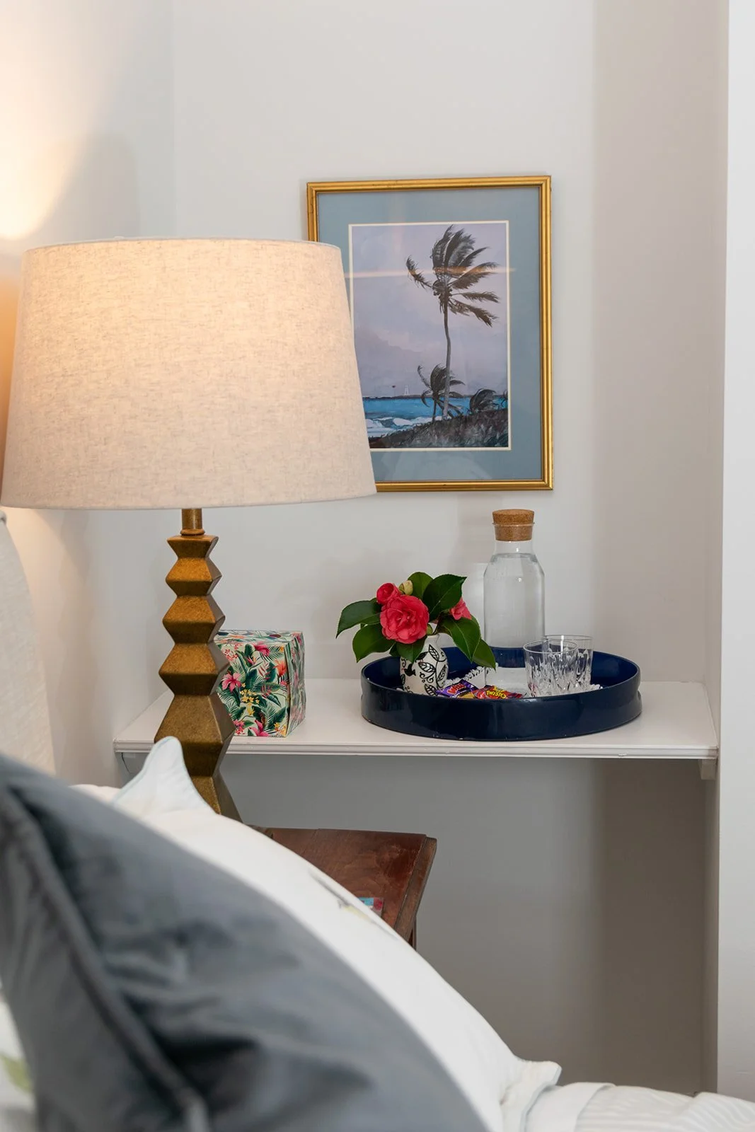 A bedside table with a floral gift box, a potted plant with pink flowers, a glass bottle with a cork, a glass, and candy. A lamp with a textured gold base and fabric shade is beside the table. A framed picture of a palm tree by the water hangs on the