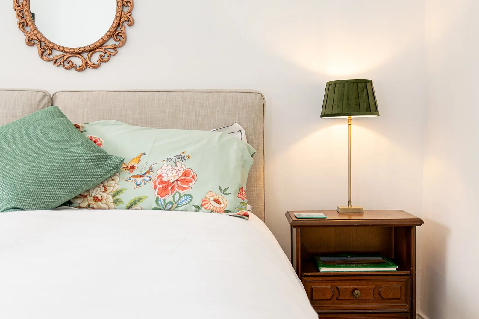 A bedroom with a bed featuring floral and solid green pillows, a beige upholstered headboard, a wooden nightstand with a green lamp, and a decorative mirror on the white wall.