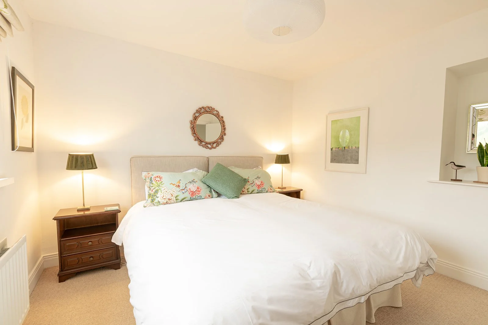 A cozy bedroom with a large bed featuring floral pillows and a green throw pillow, bedside tables with lamps, a round ornate mirror above the headboard, abstract artwork on the walls, a window with a plant and decorative bird, and soft lighting creat