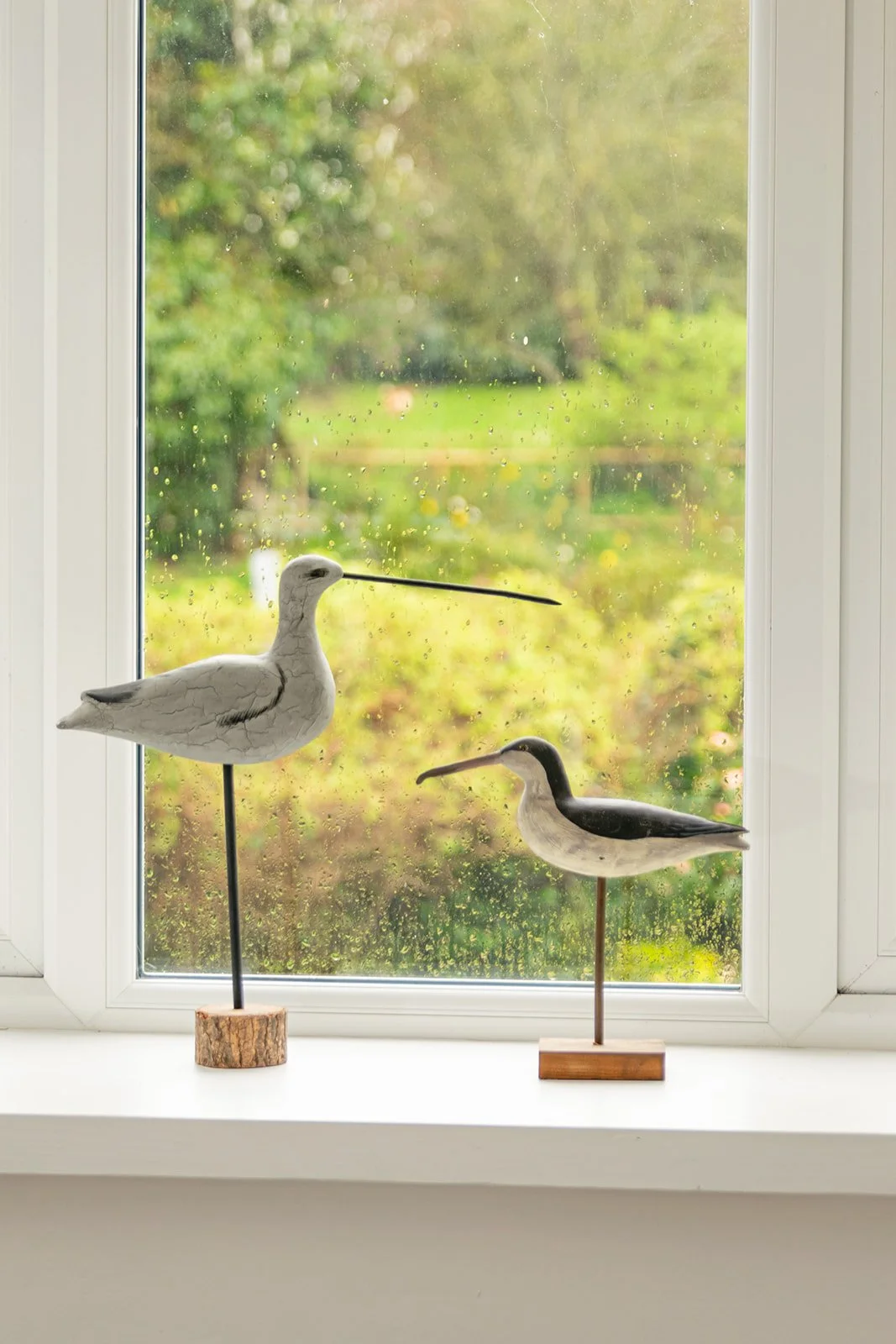 Decorative bird figures on window sill with a window showing a green garden outside.