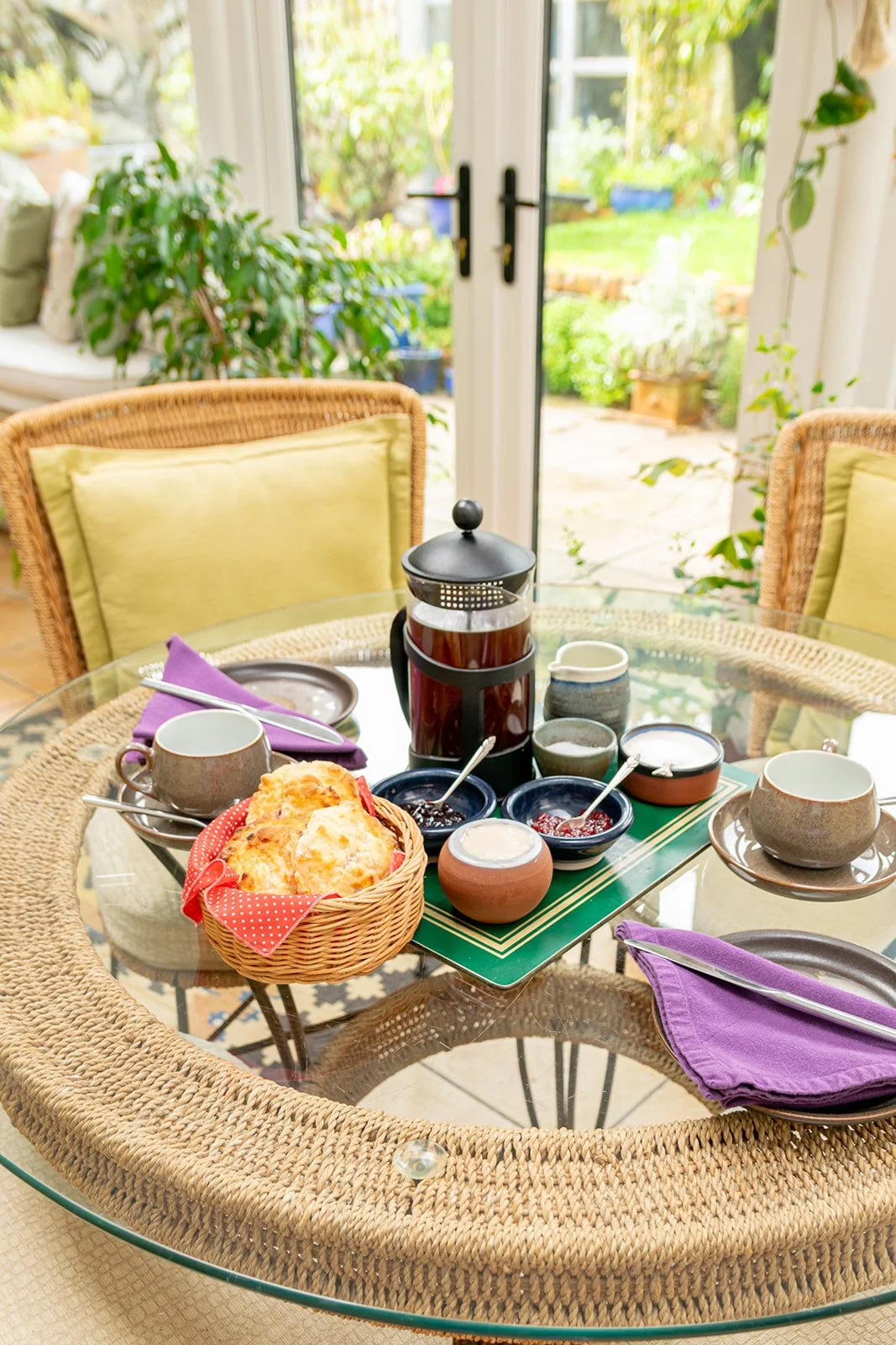 Breakfast table set with scones, jam, coffee, tea cups, and utensils in a sunlit indoor patio with greenery outside.