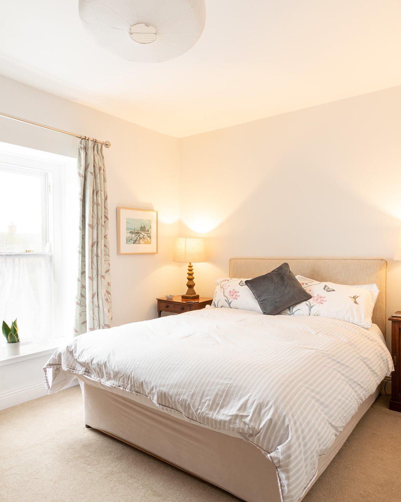 A neatly made bed with white sheets, floral pillowcases, and a dark gray throw pillow in a bright bedroom. There is a wooden nightstand with a lamp beside the bed and a framed picture on the wall. A window with patterned curtains is partially visible, with some plants on the windowsill.