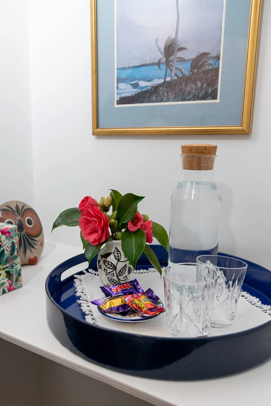 A white tray with a white doily, two glasses, a glass water bottle with a cork, a small potted pink camellia flower, and three wrapped candies on a table. In the background, a framed painting of a tropical scene with palm trees and ocean waves hangs 