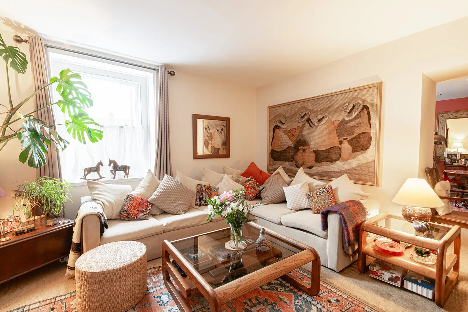 A cozy living room with a sectional sofa, decorative pillows, a coffee table with a glass top, and various plants. There is a large window with curtains, and paintings on the wall. A side table with a lamp and decorations is nearby.