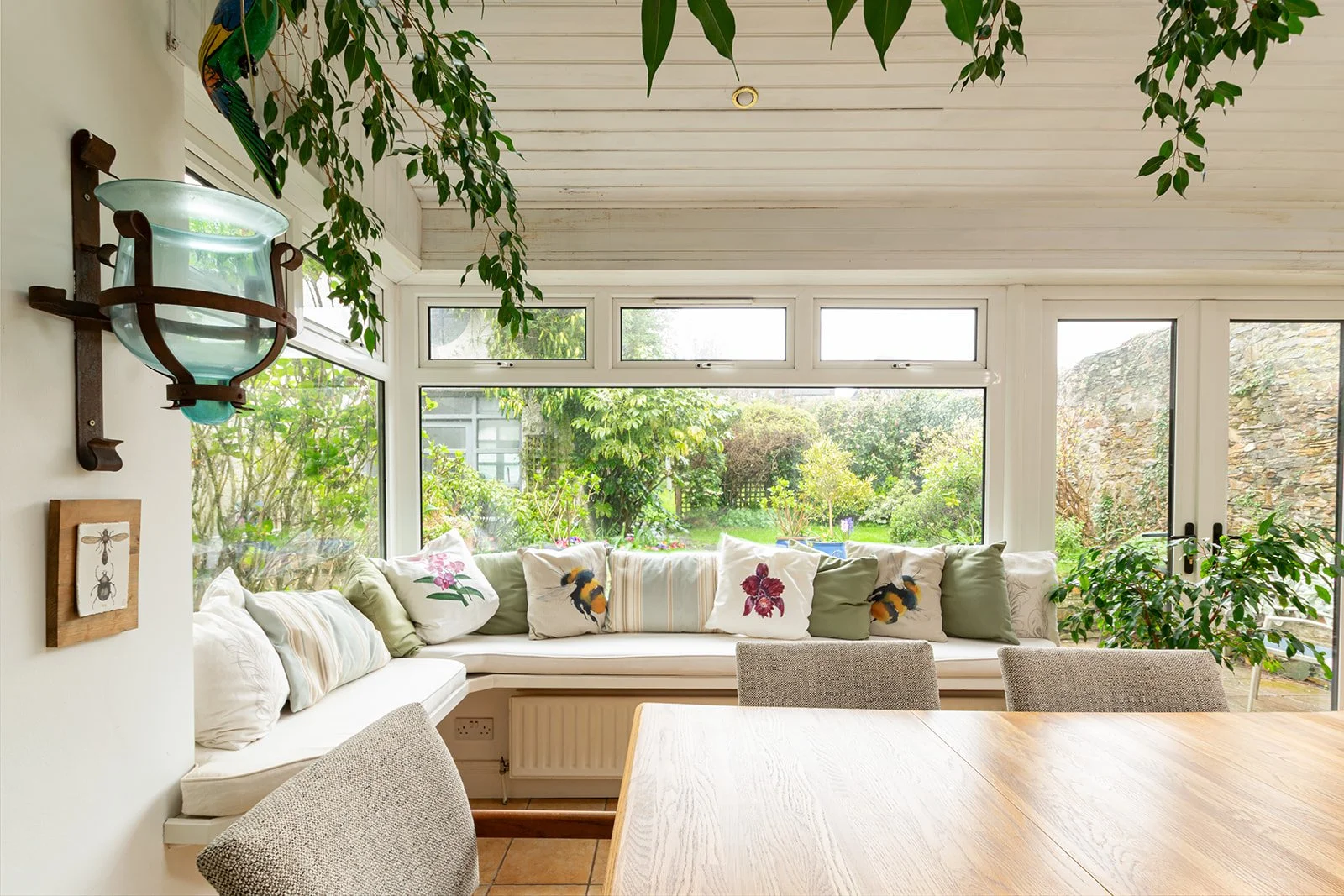 Sunroom with white cushioned seating area decorated with pillows featuring floral and insect designs, surrounded by large windows overlooking a lush green garden, and accented with hanging plants and a wall-mounted decorative glass lamp.