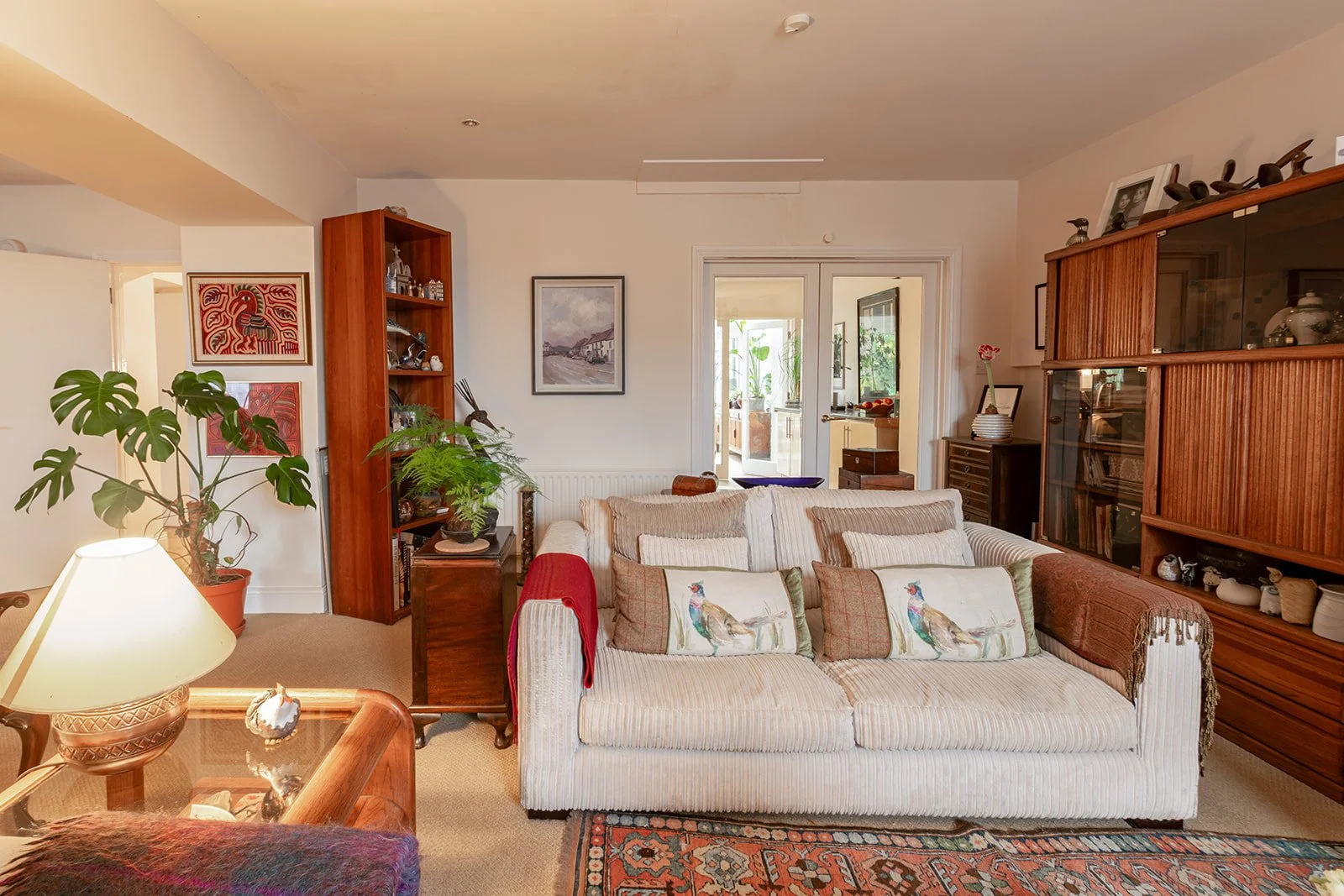 A cozy living room with a white, striped sofa decorated with bird-themed pillows, wooden furniture, a large green houseplant, and artwork on the walls.