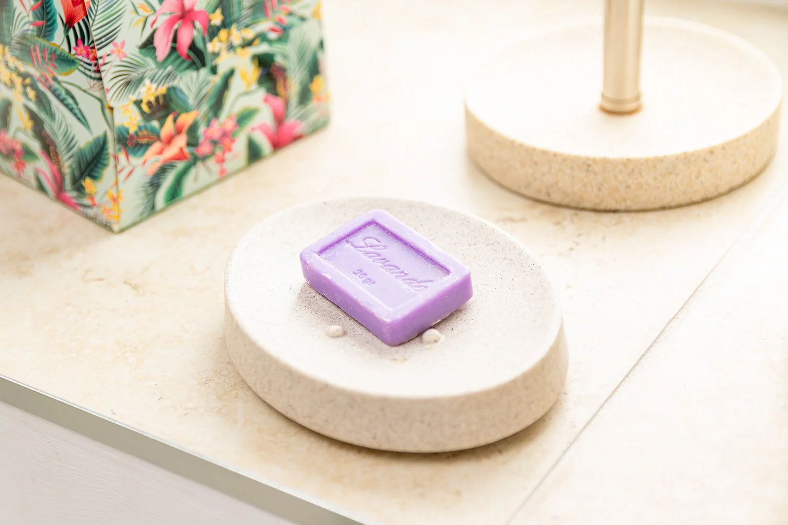 Purple soap labeled 'Glamour 2022' on a round, beige stone display stand with a small white decorative item, with a tropical floral gift bag and a beige table lamp in the background.