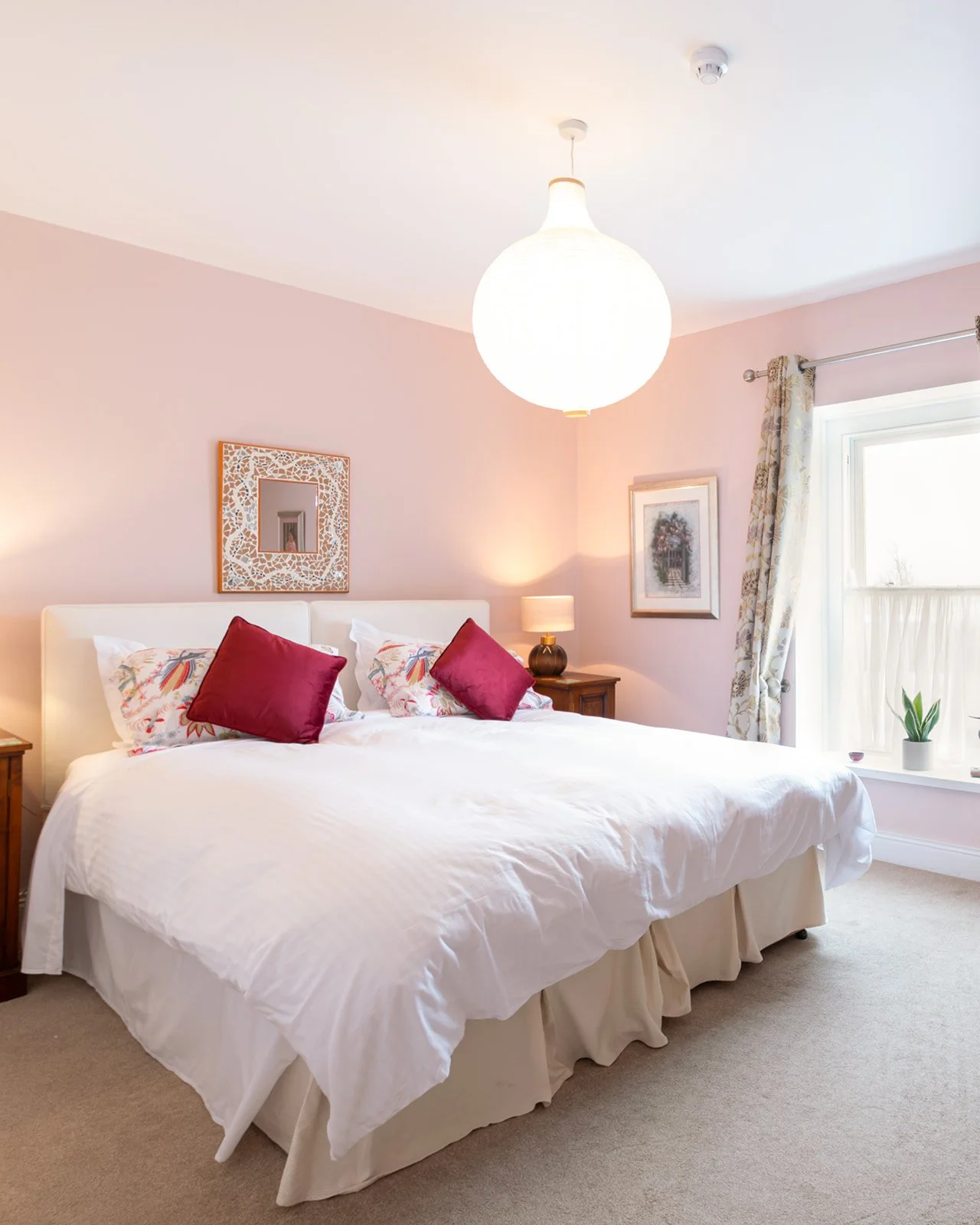 A bedroom with a large bed covered in white bedding, two red pillows, and decorative pillows with floral patterns. There is a wooden nightstand with a lamp, framed artwork, a window with floral curtains, and a small potted plant on the windowsill.