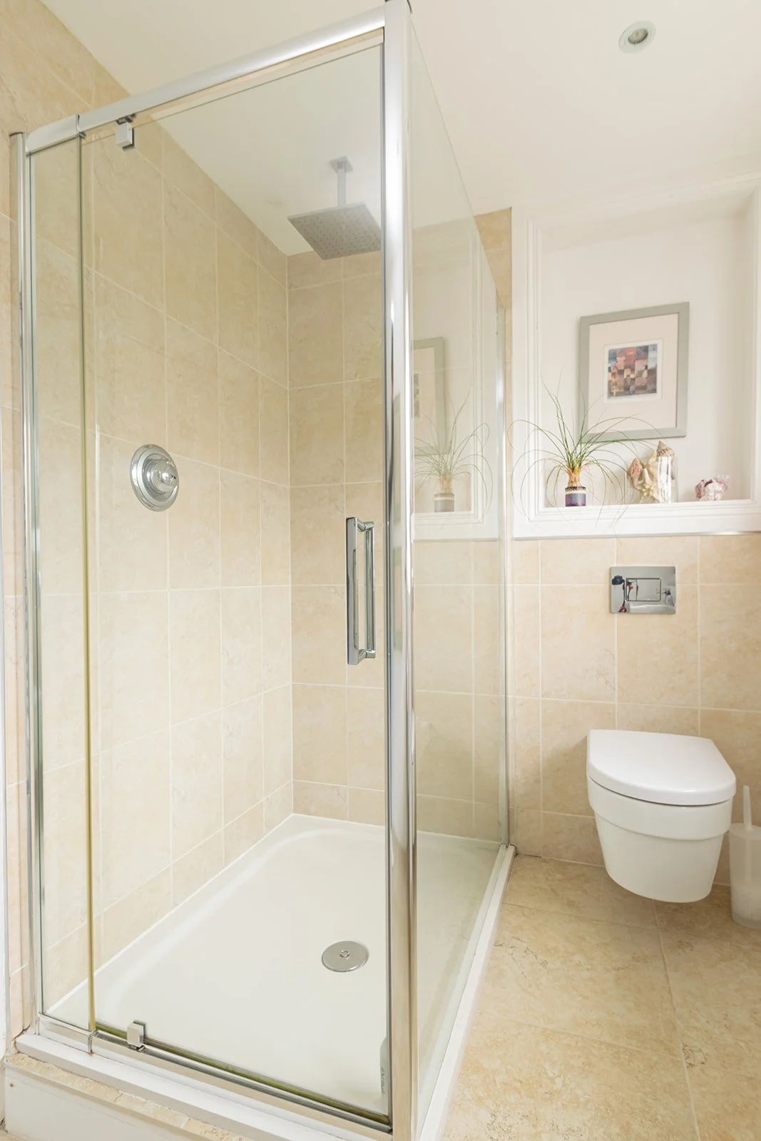 Bathroom with glass-enclosed shower and wall-mounted toilet, beige tiled walls and floor, decorative plants and framed art on window ledge, and a small recessed light in ceiling.