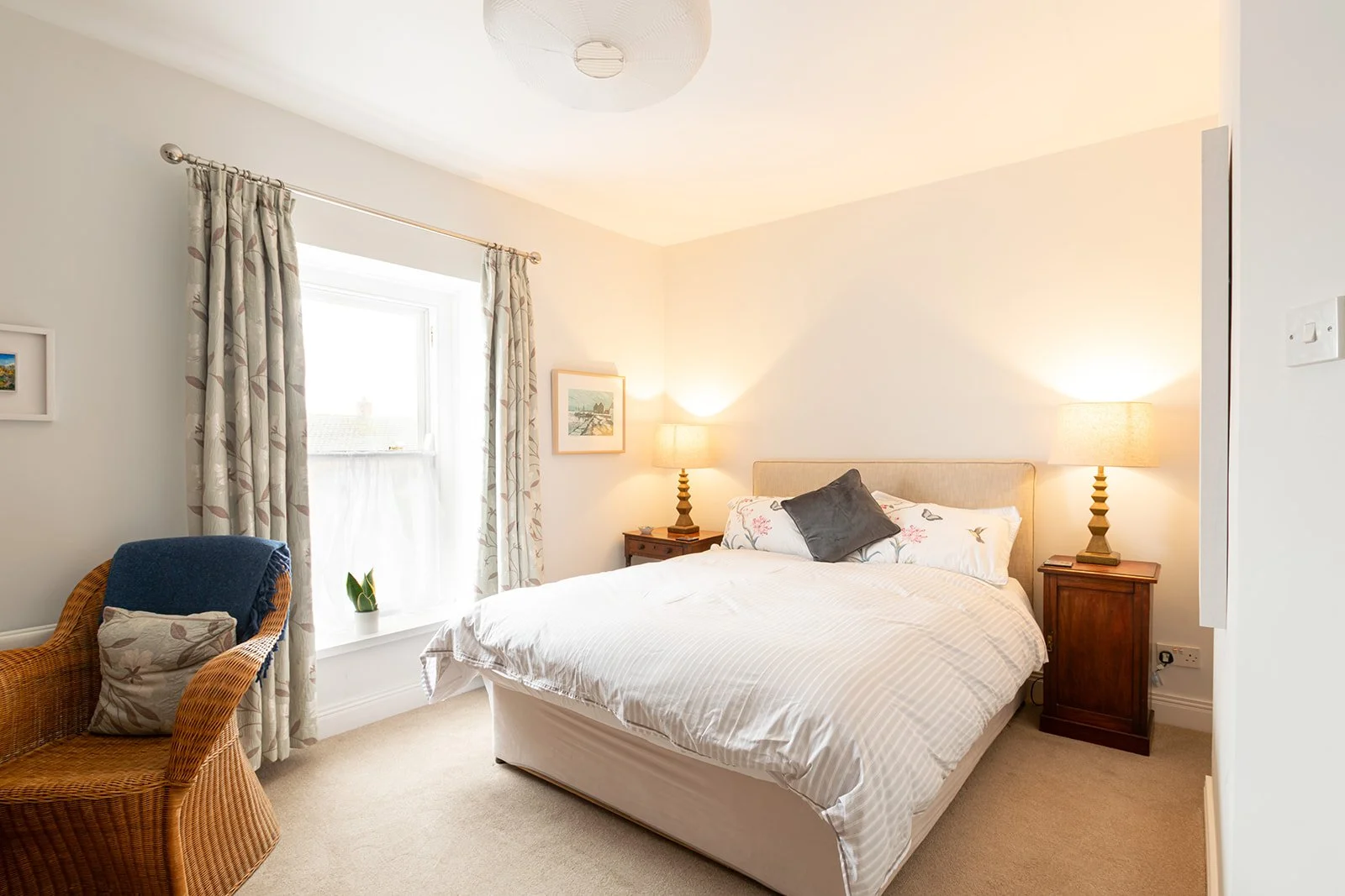 A cozy bedroom with a bed, two nightstands with lamps, a window with curtains, a wicker chair with a pillow, and artwork on the walls.