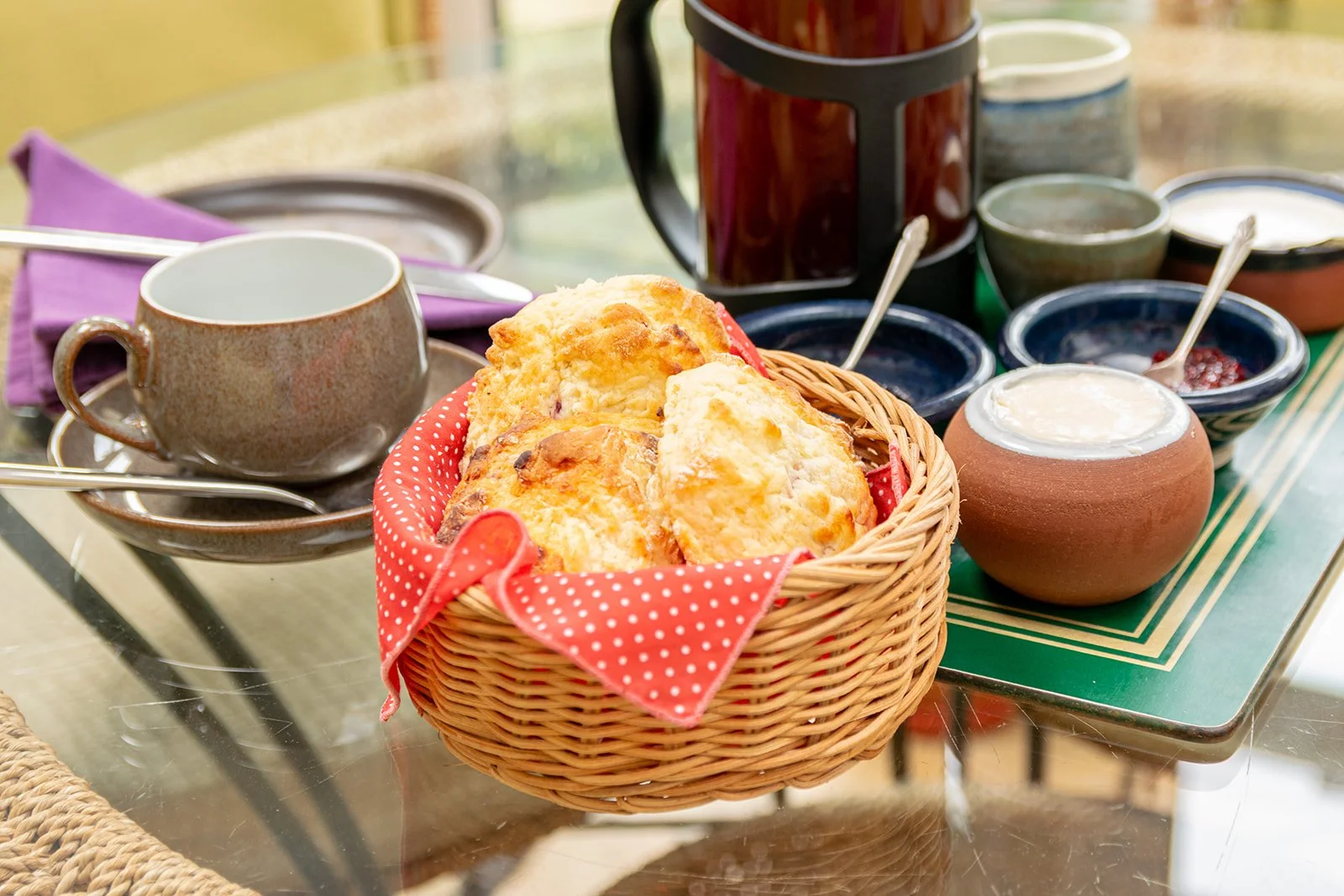 A wicker basket filled with scones, placed on a glass table with tea cups, saucers, jars of jam, and a teapot, set for breakfast.
