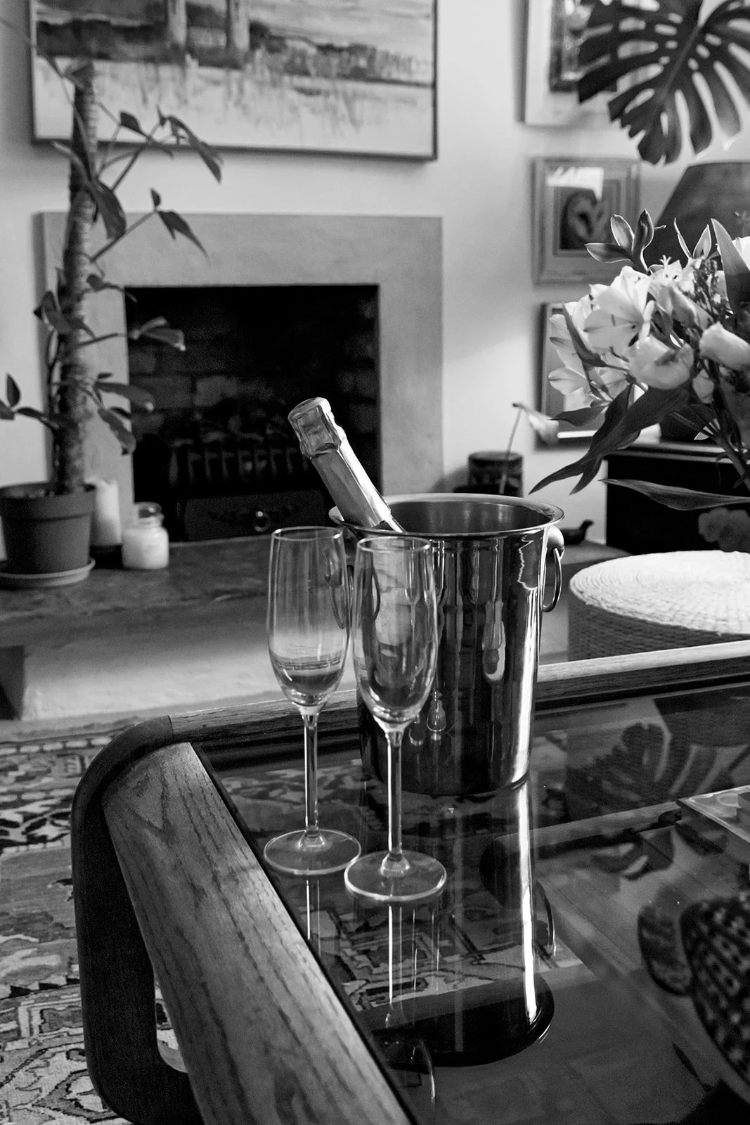 A table with two champagne flutes, a champagne bottle in an ice bucket, and a plant on a wooden table inside a living room with artwork and plants in the background.