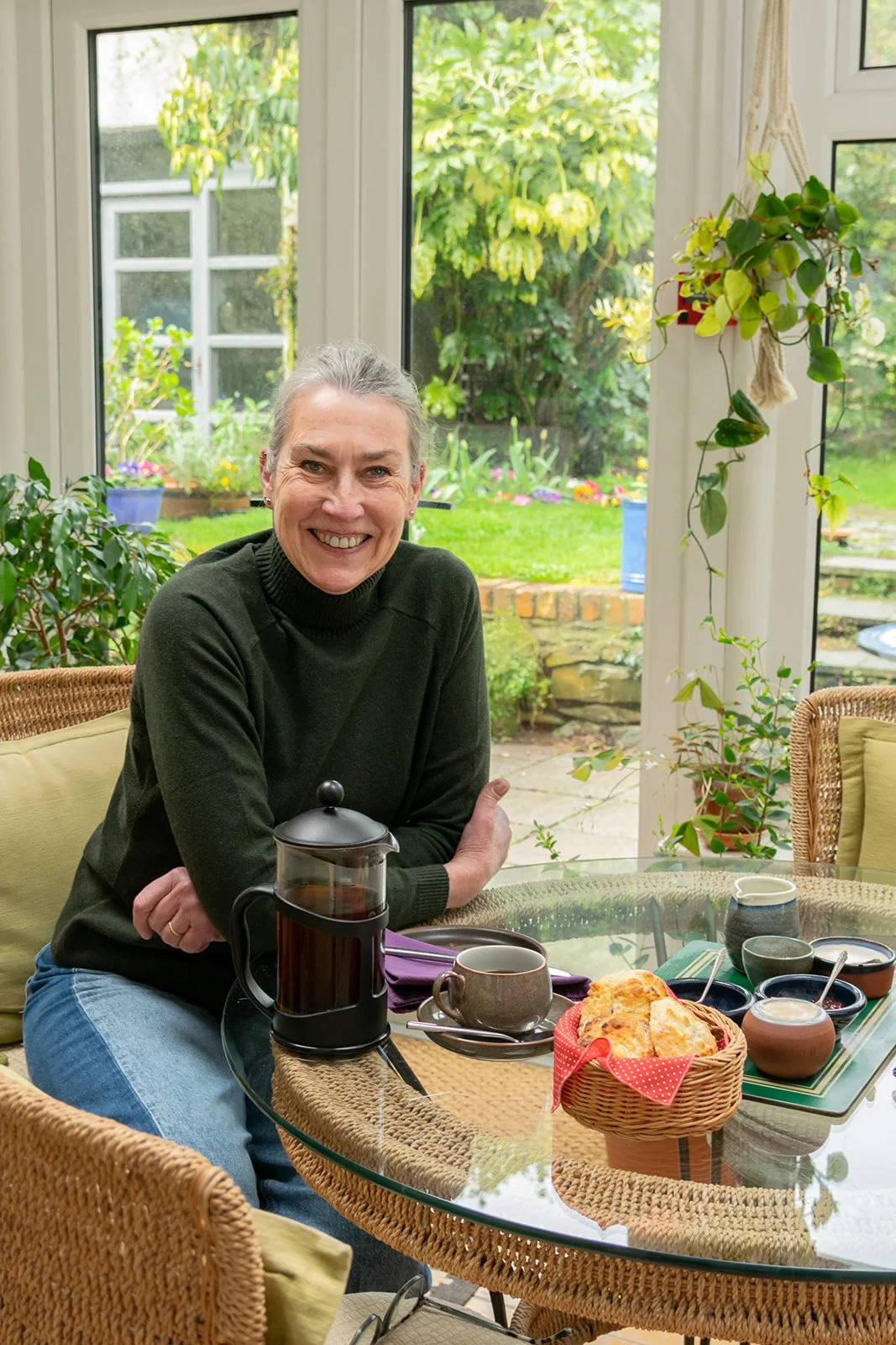 A smiling woman with gray hair sitting at a glass-topped wicker table in a sunlit room with large windows. The table has a French press with coffee, a cup, and a basket of scones. There are various small bowls and cups on the table, and a garden with green plants and flowers outside.