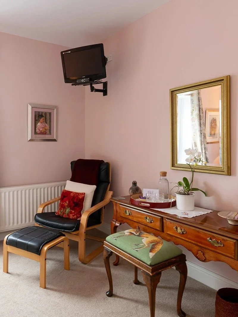 Living room corner with a wall-mounted TV, framed paintings, a leather and wood armchair with a pillow, a wooden side table with decorative items and a potted orchid, a upholstered stool with a fox print, and a beige carpet.