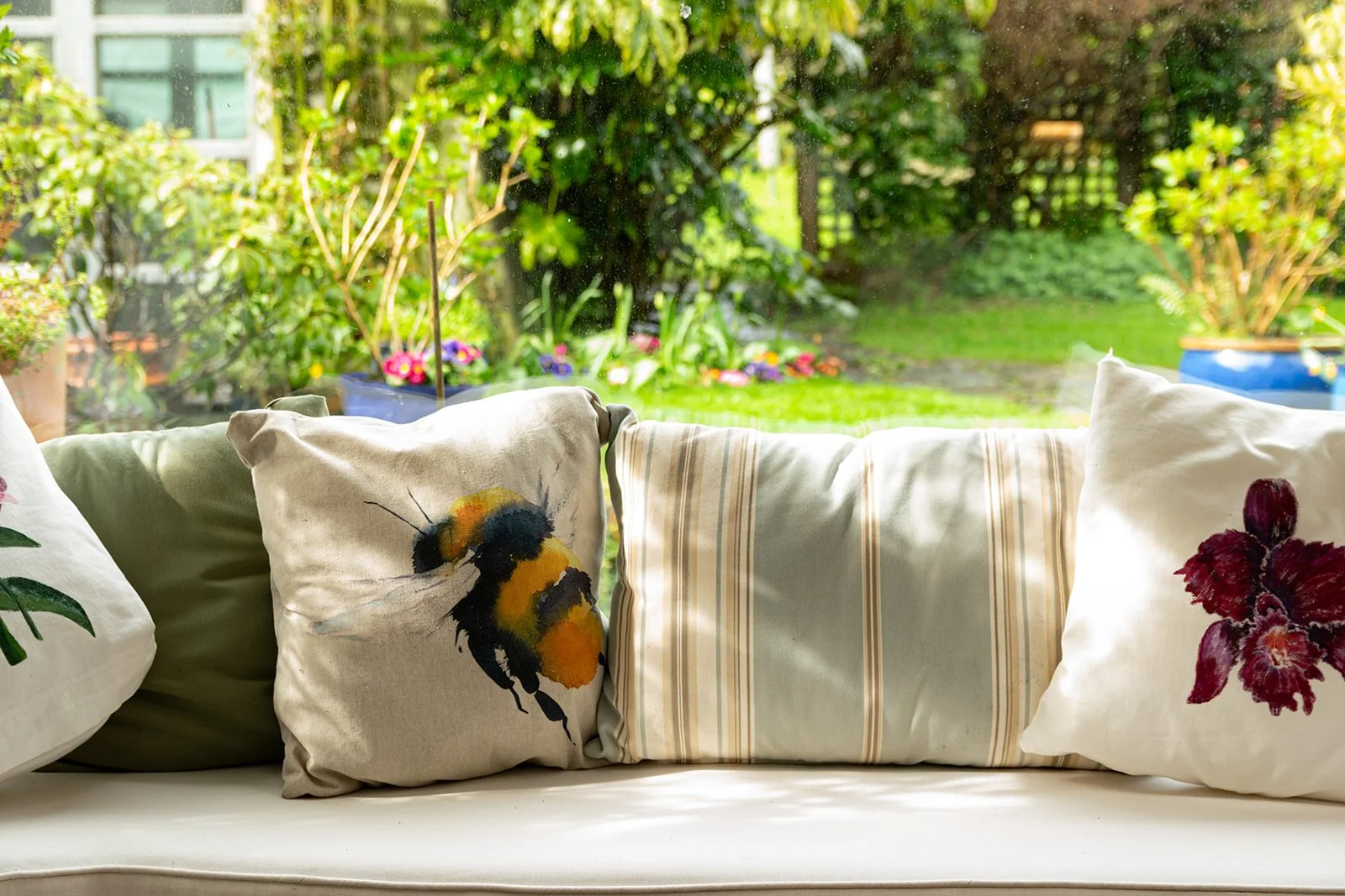 A bright, airy room with a sofa adorned with decorative pillows featuring floral and bee designs, overlooking a lush garden with green plants, flowers, and a wooden fence outside.