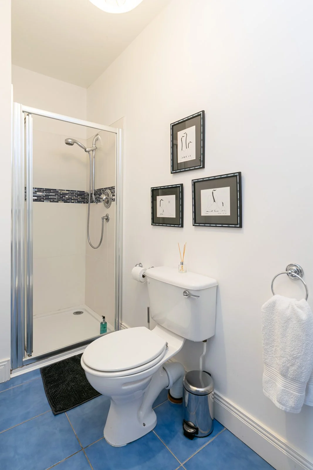 A small bathroom with a shower stall, white toilet, black rug, blue tiled floor, wall art, towel, and toilet brush.