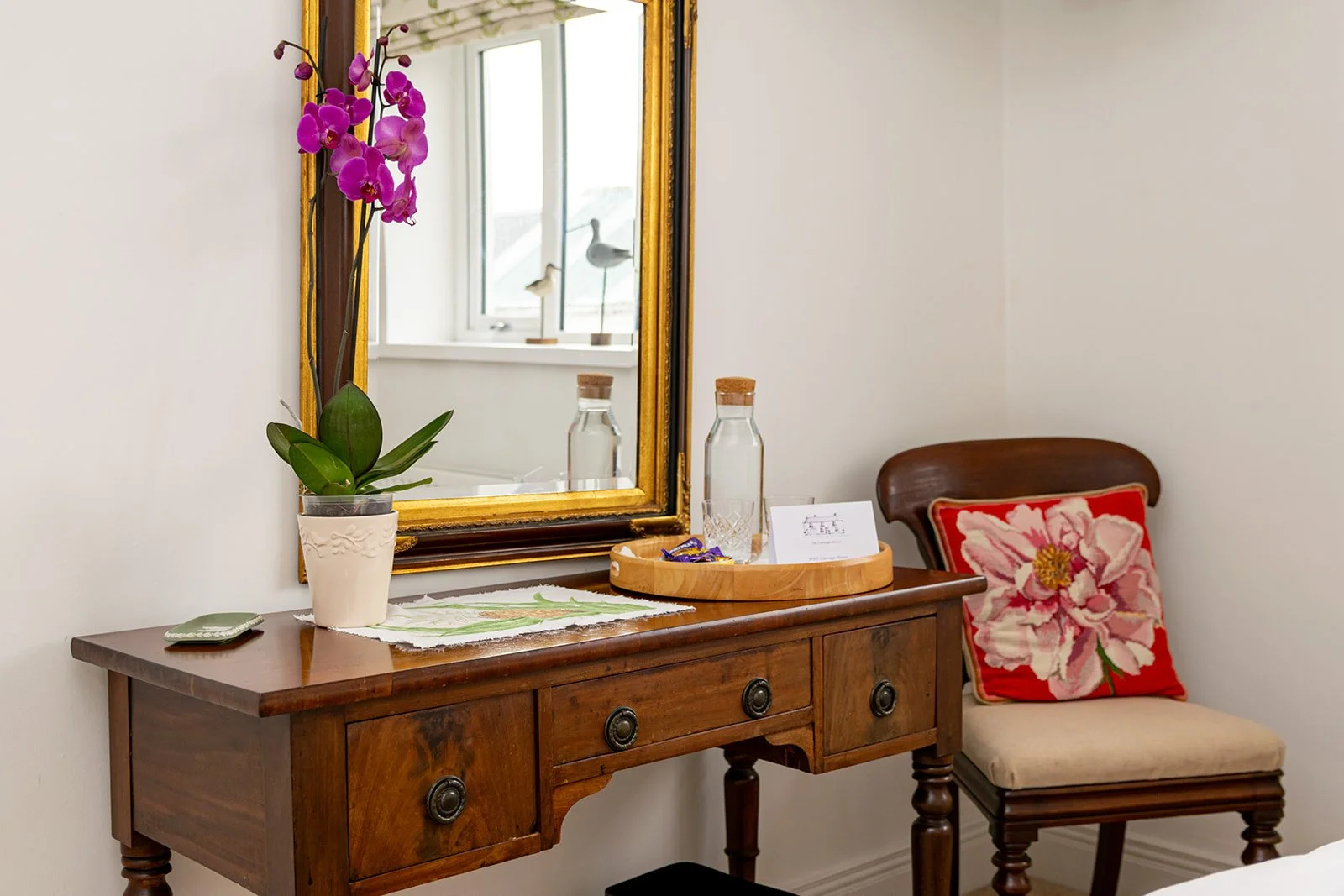 A wooden dresser with a mirror, decorated with a potted orchid plant, two glass bottles with cork stoppers, a tray with purple wrapped candies, and a card. Beside the dresser, a wooden chair with a floral cushion features a large pink flower on a red background. The mirror reflects window views with bird figurines on the windowsill.