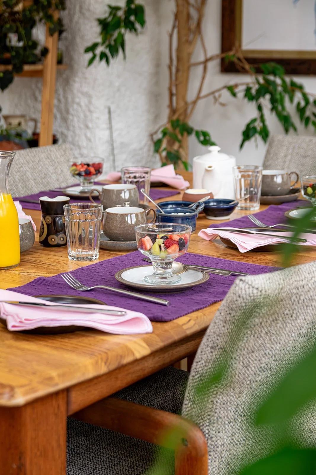 A wooden dining table set for breakfast or brunch with purple placemats, pink napkins, clear glasses, coffee cups, utensils, a bowl of mixed fruit, a jug of orange juice, and a white teapot, surrounded by grey upholstered chairs, in a cozy room with 