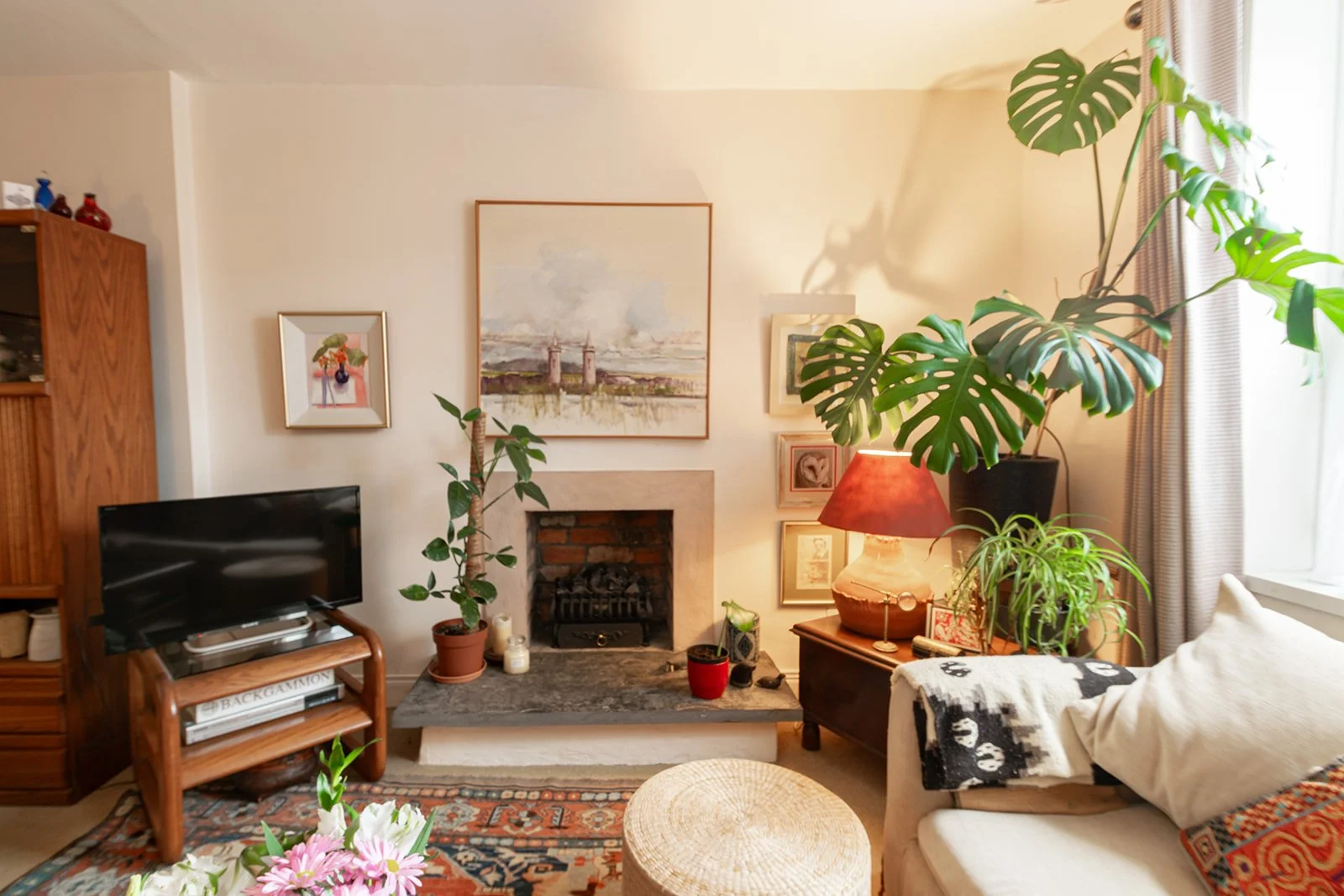 Living room with a fireplace, a small TV on a wooden stand, several potted plants, a table lamp with a red shade, artwork on the wall, and a white couch with a blanket and cushion near a window.