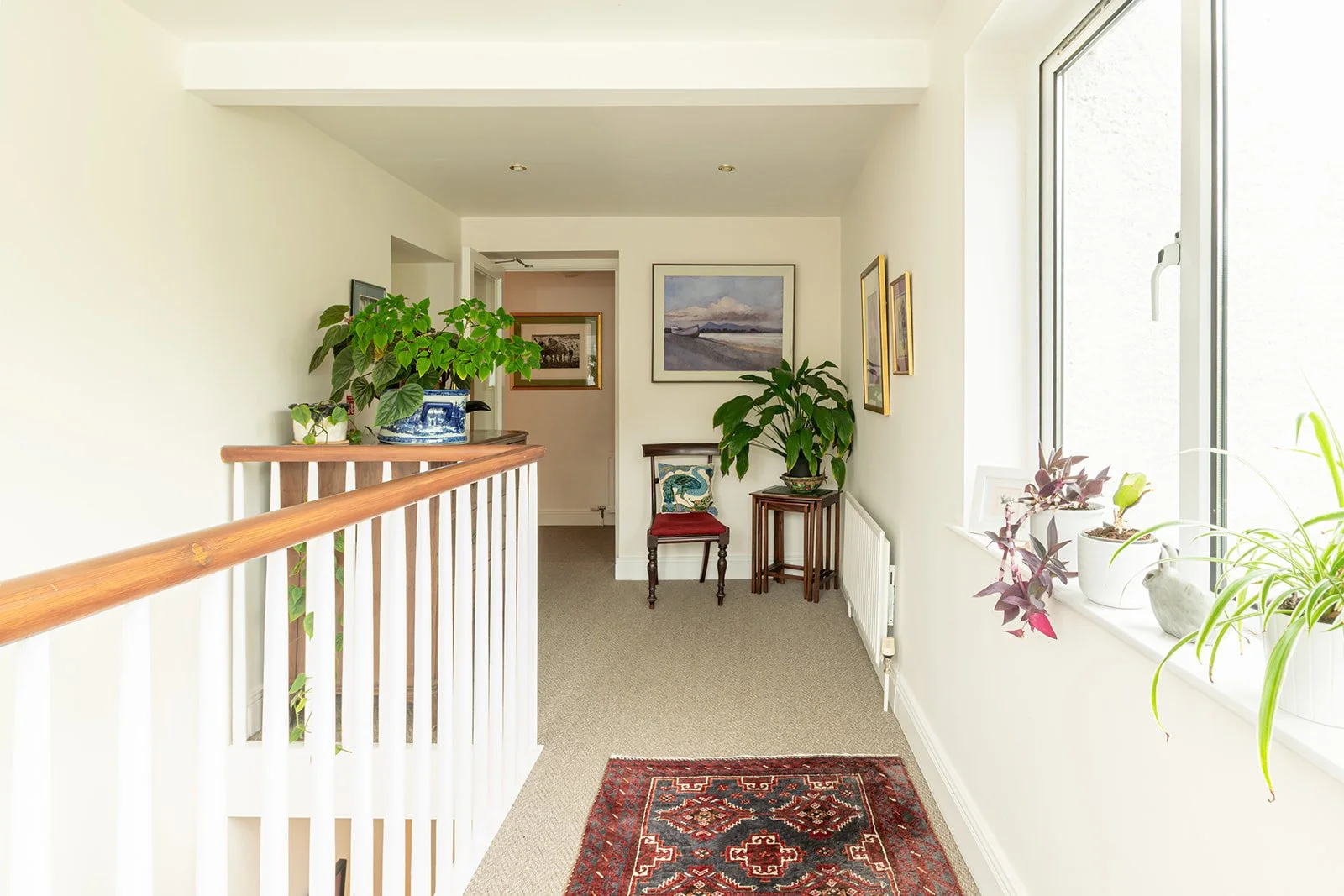 Interior view of a bright hallway with plants on the windowsill and side tables, framed artwork on the walls, and a decorative rug on beige carpet.