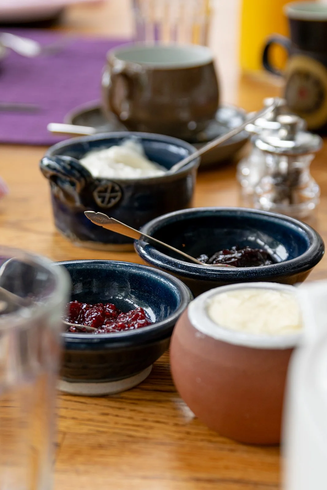 Various jars of jam and cream on a wooden table, with a purple placemat and some blurred background objects.