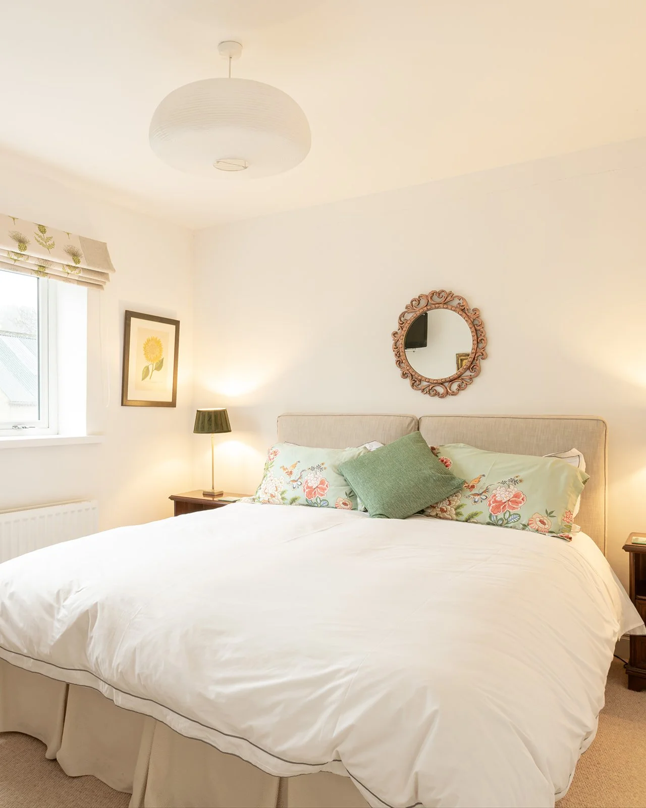 A cozy bedroom with a neatly made bed featuring floral-patterned pillows and a green accent pillow, illuminated by soft lighting from a bedside lamp. The room has a window with a decorative blind, a framed floral picture on the wall, a round mirror above the bed, and neutral-toned furnishings.