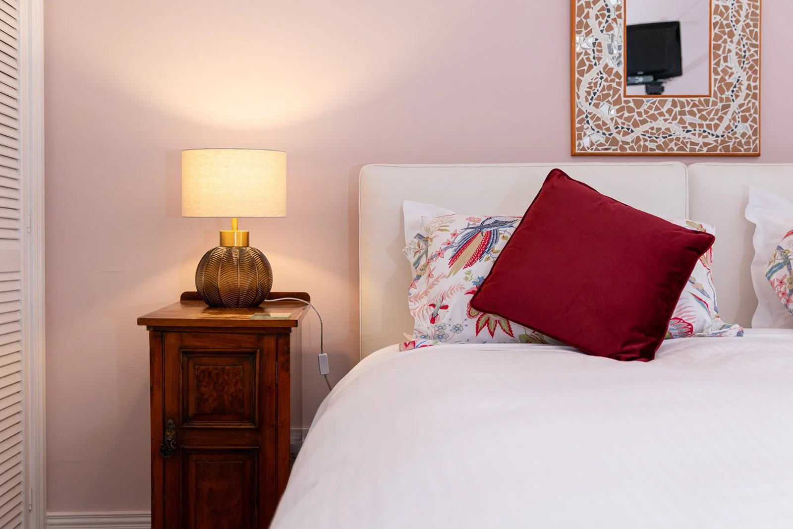 A bedroom with a bedside table, a lamp, a floral pillow, a red cushion, and a mirror on the wall.