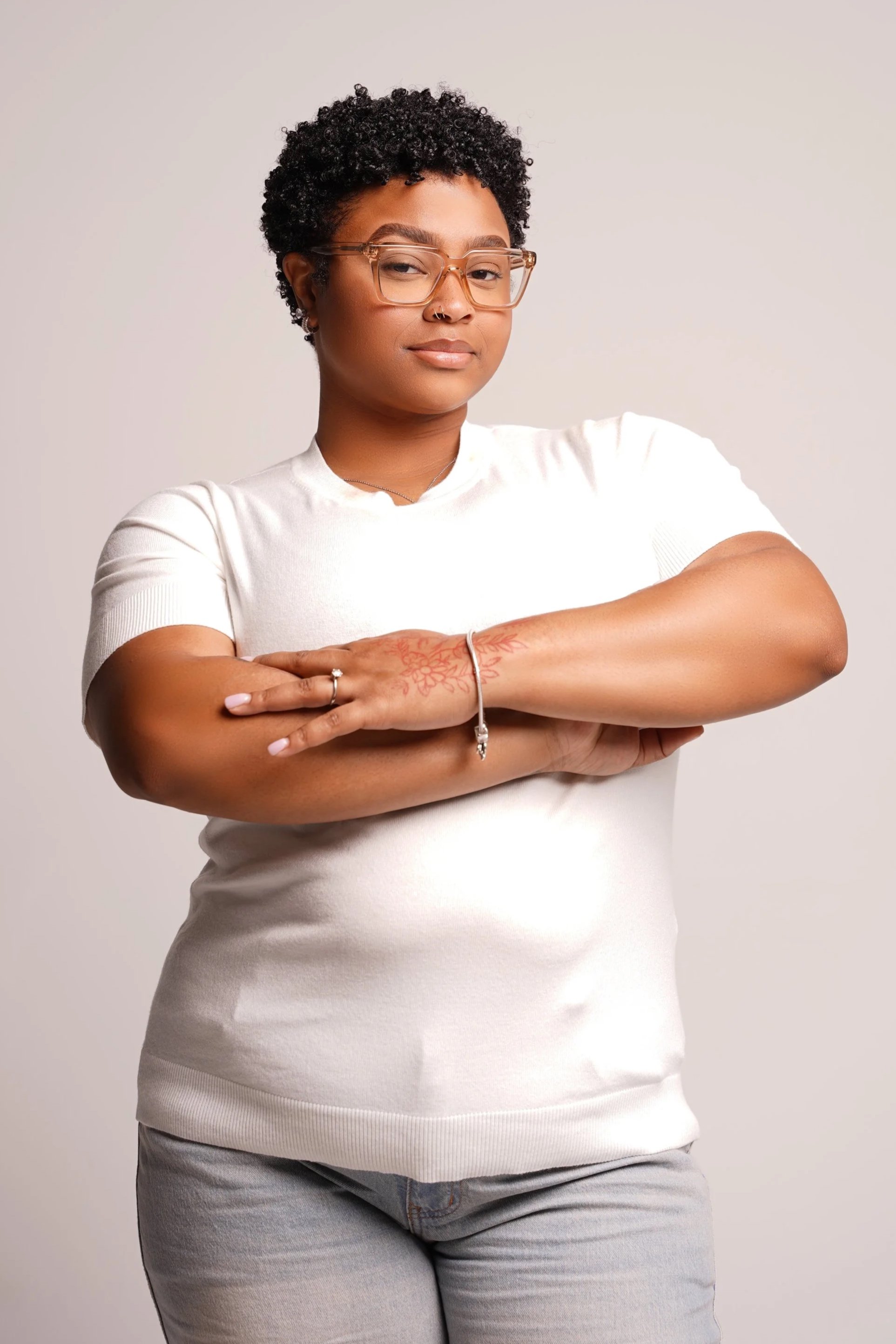 A woman with short curly hair wearing glasses and a white t-shirt, crossing her arms and looking confidently at the camera.