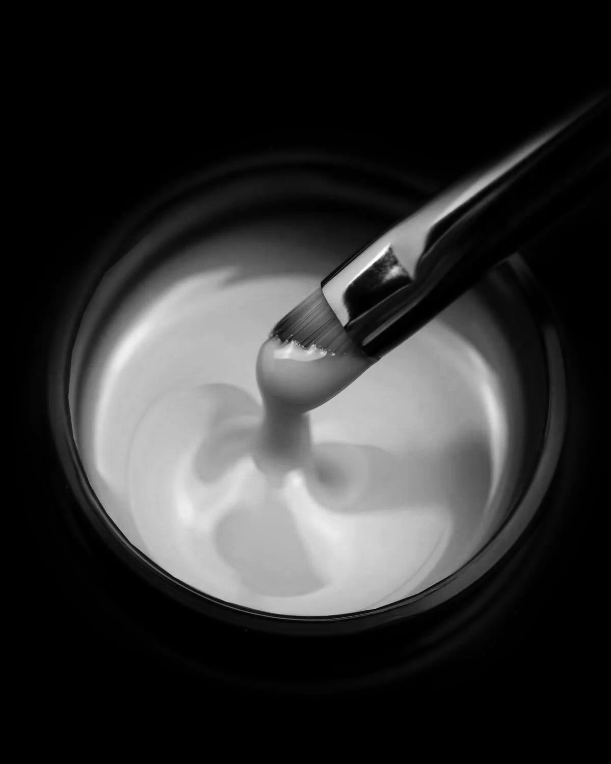 A close-up black and white image of a makeup brush swirling in a container of cream or lotion.