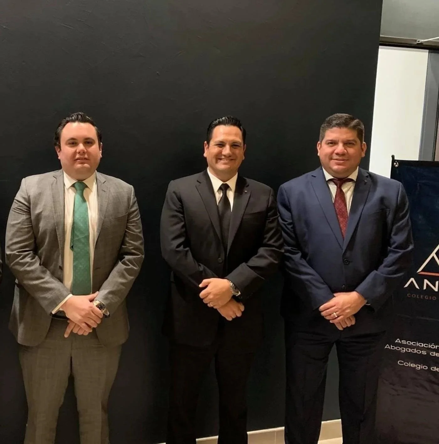 Tres hombres con trajes formales posando para una foto frente a un fondo negro con un cartel que dice 'Asociación de Abogados'.