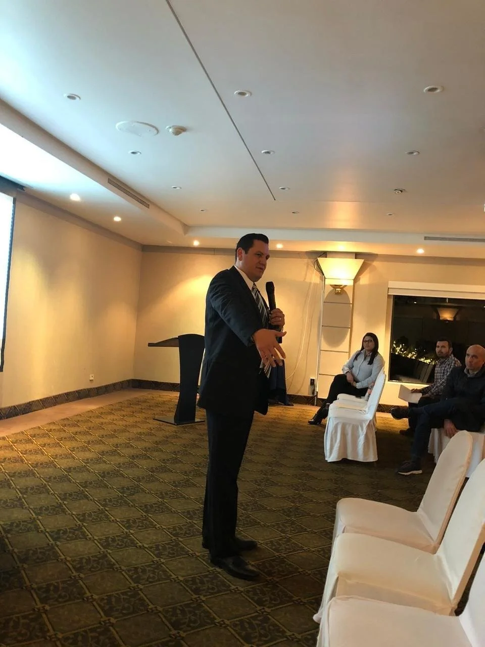 hombre en traje hablando con micrófono en una sala de reuniones, con varias personas sentadas escuchando, en un ambiente formal y bien iluminado