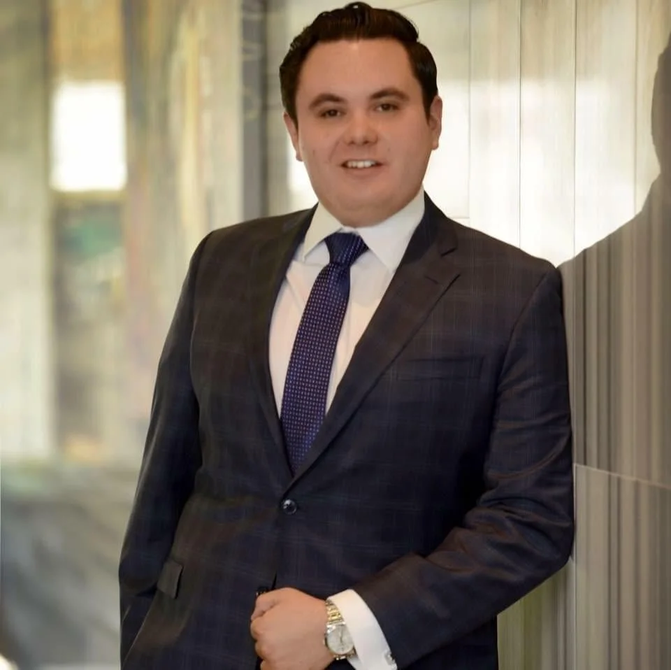 Hombre vestido con traje oscuro, camisa blanca y corbata azul, posando en un pasillo con paredes de madera.