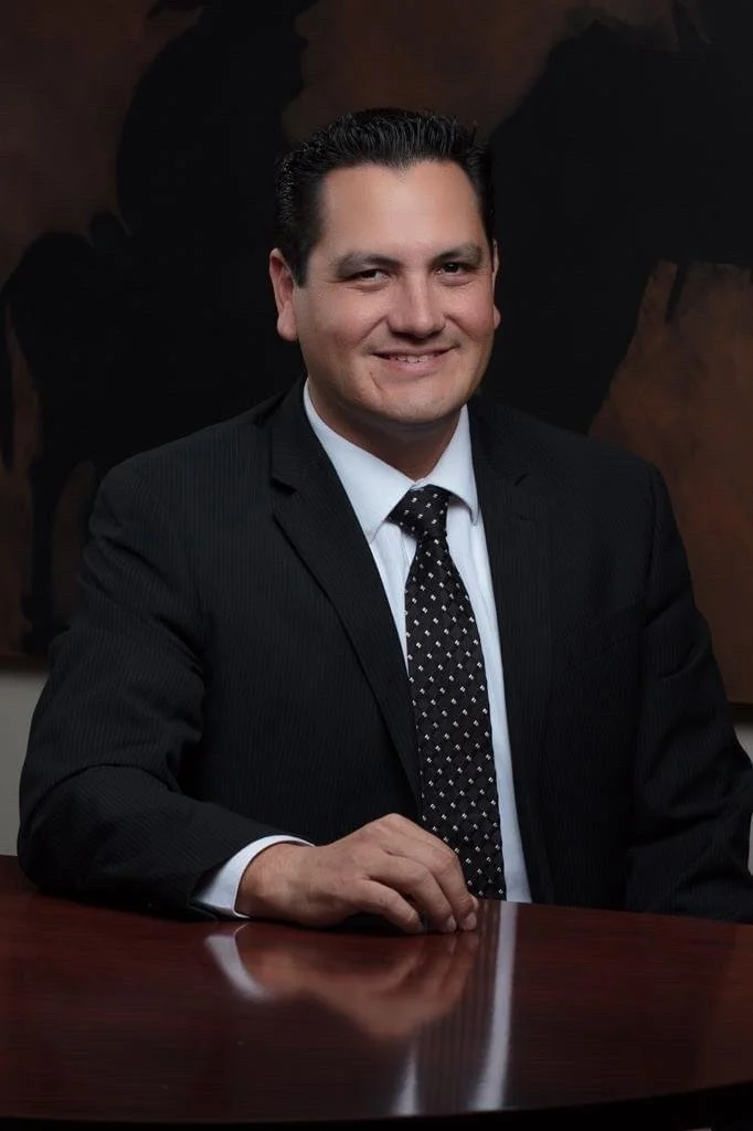 Retrato de un hombre con saco negro, camisa blanca y corbata con puntos blancos, sentado frente a una mesa de madera en un ambiente profesional.