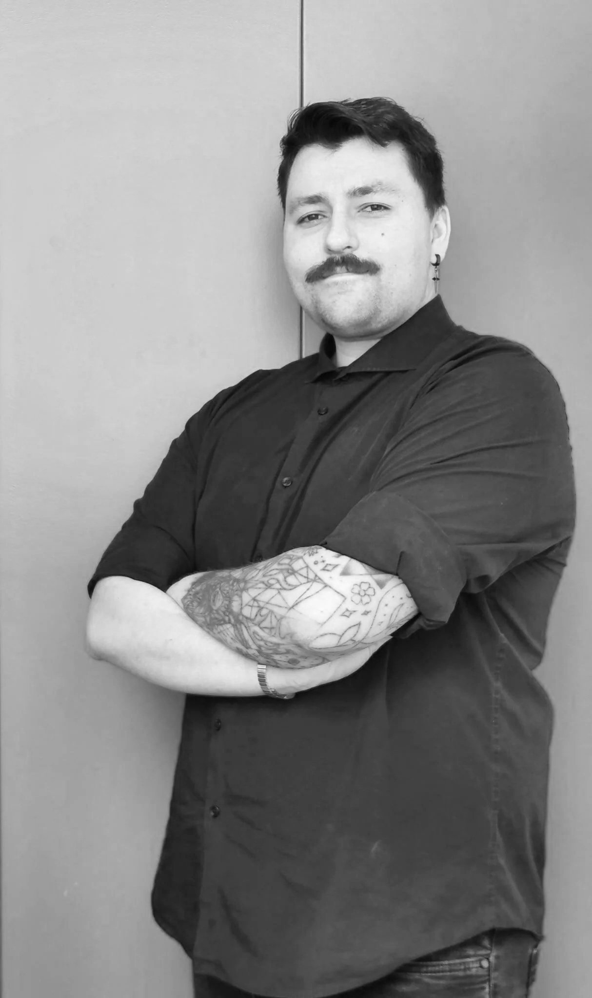 Black and white photo of a man with dark hair, mustache, and earrings, crossing his arms and smiling slightly, wearing a dark button-up shirt, standing against a plain wall.