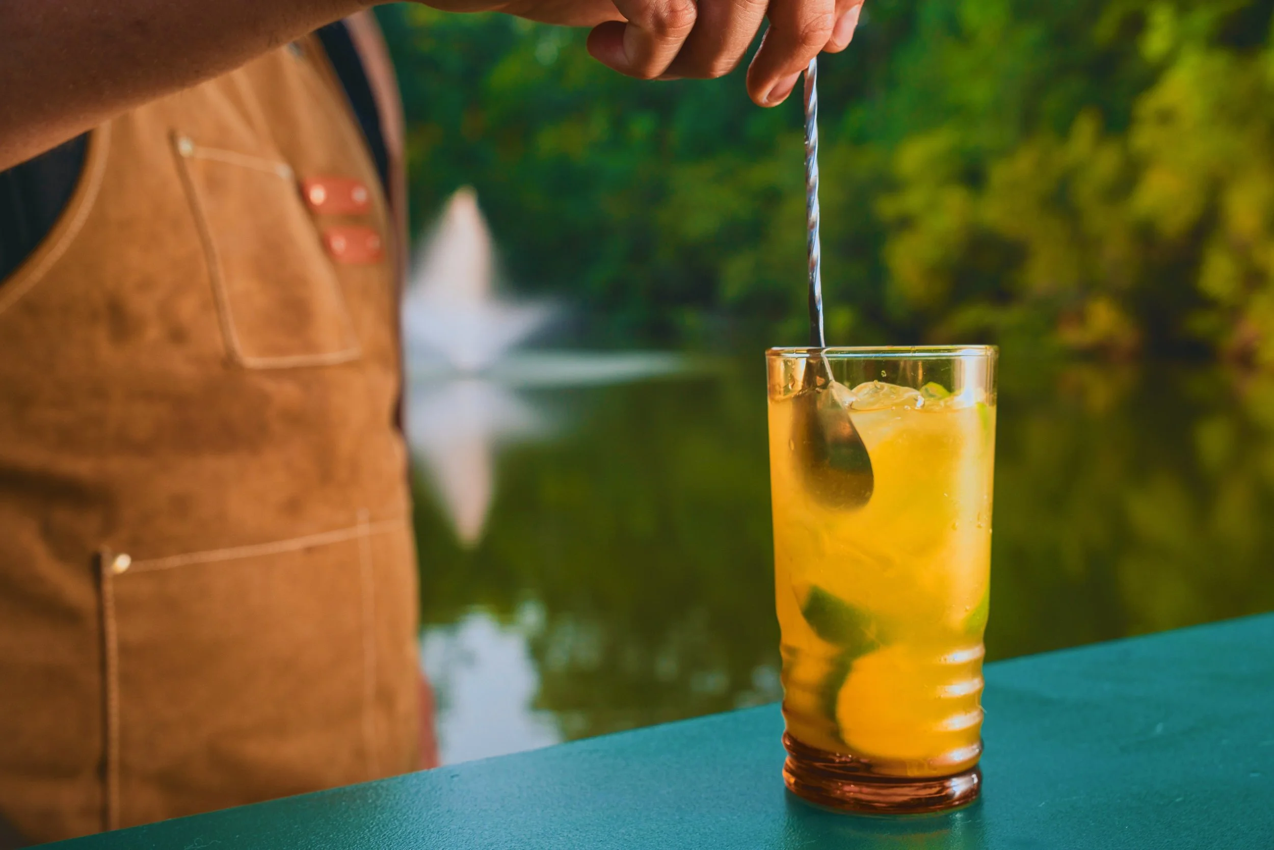 A person is pouring a drink from a bottle into a tall glass with ice and lime slices outdoors with green trees in the background.