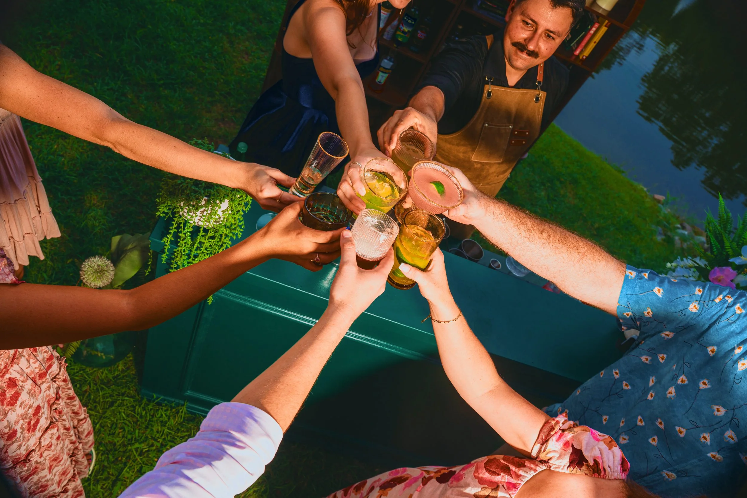 Group of friends toasting drinks outdoors near a body of water, with a person wearing an apron serving drinks.