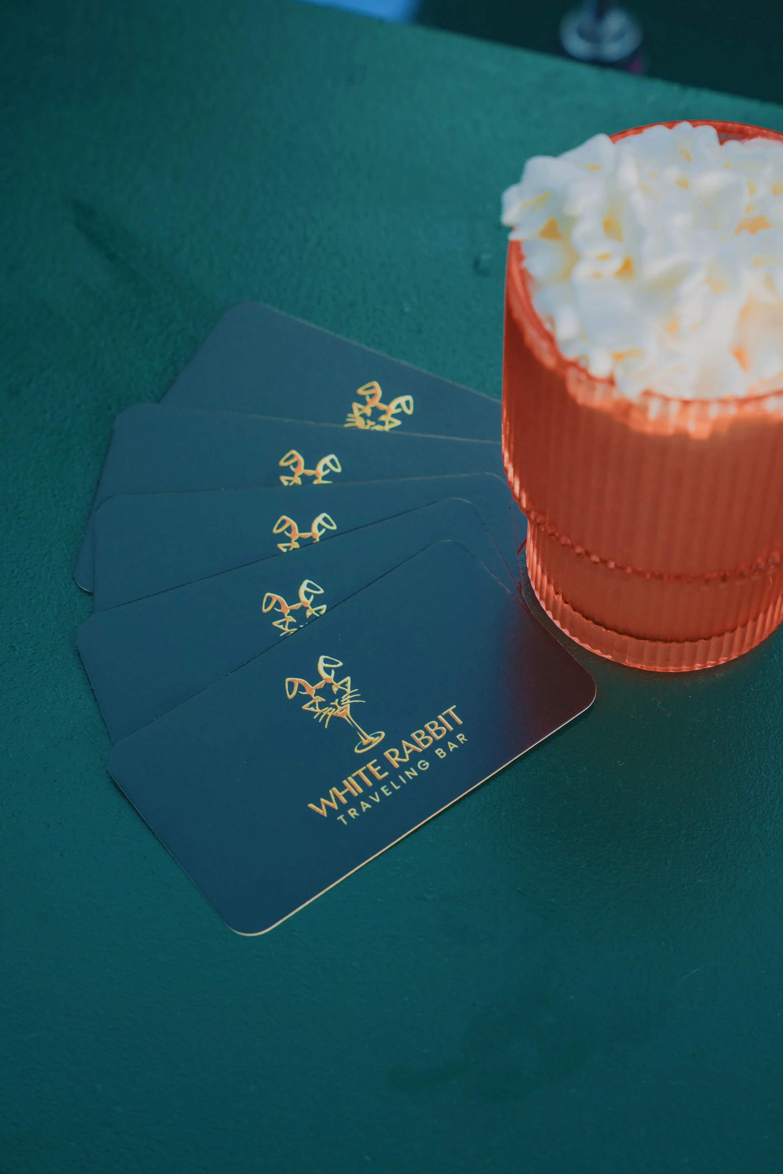 Seven blue loyalty or membership cards with a golden rabbit logo and the text 'White Rabbit Traveling Bar' on a turquoise surface, next to an orange cup filled with whipped cream.