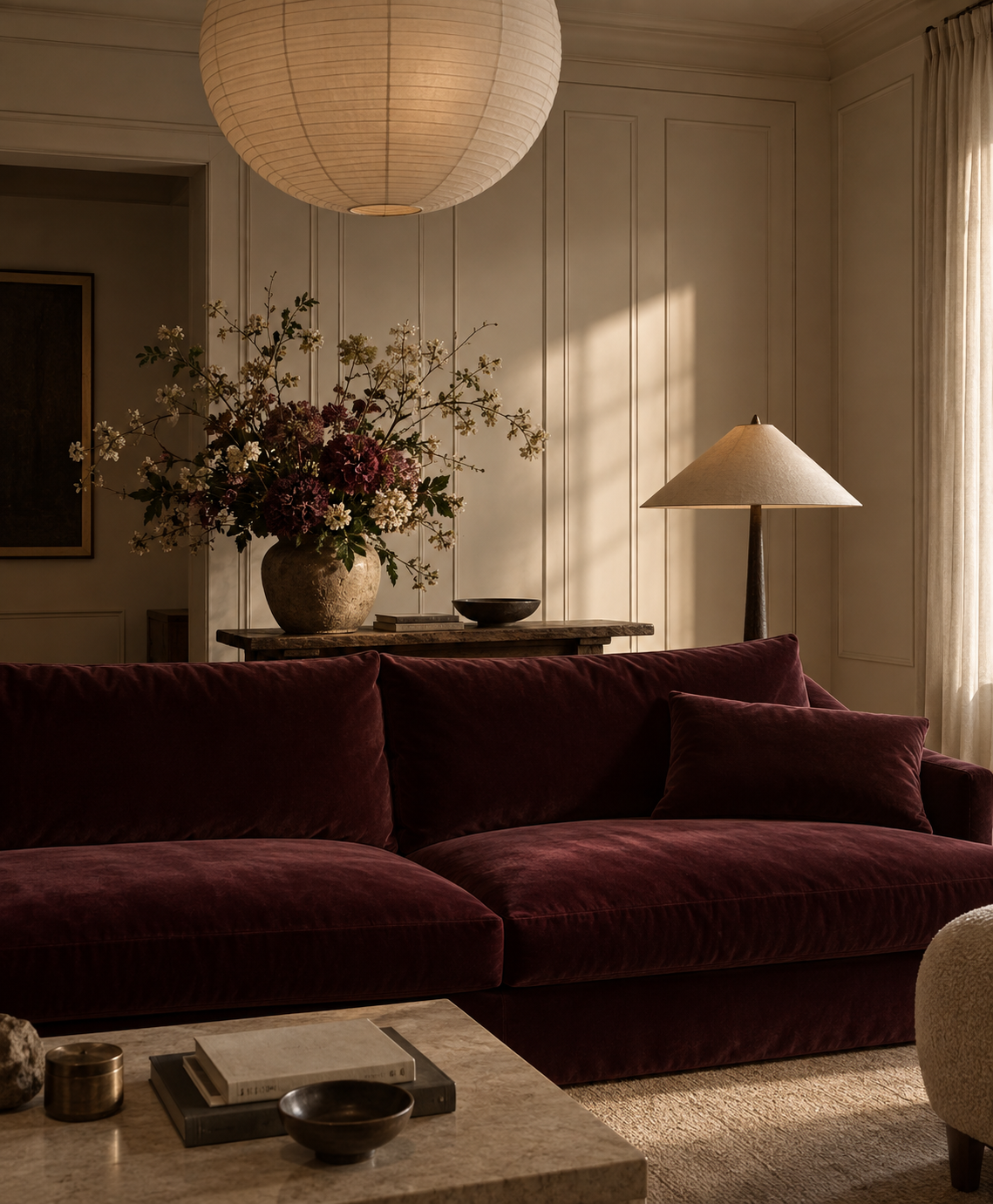 Interior of a cozy living room with a dark red velvet sofa, a marble coffee table with books and bowls, a large floral arrangement, a floor lamp, and natural light coming through a window with curtains.