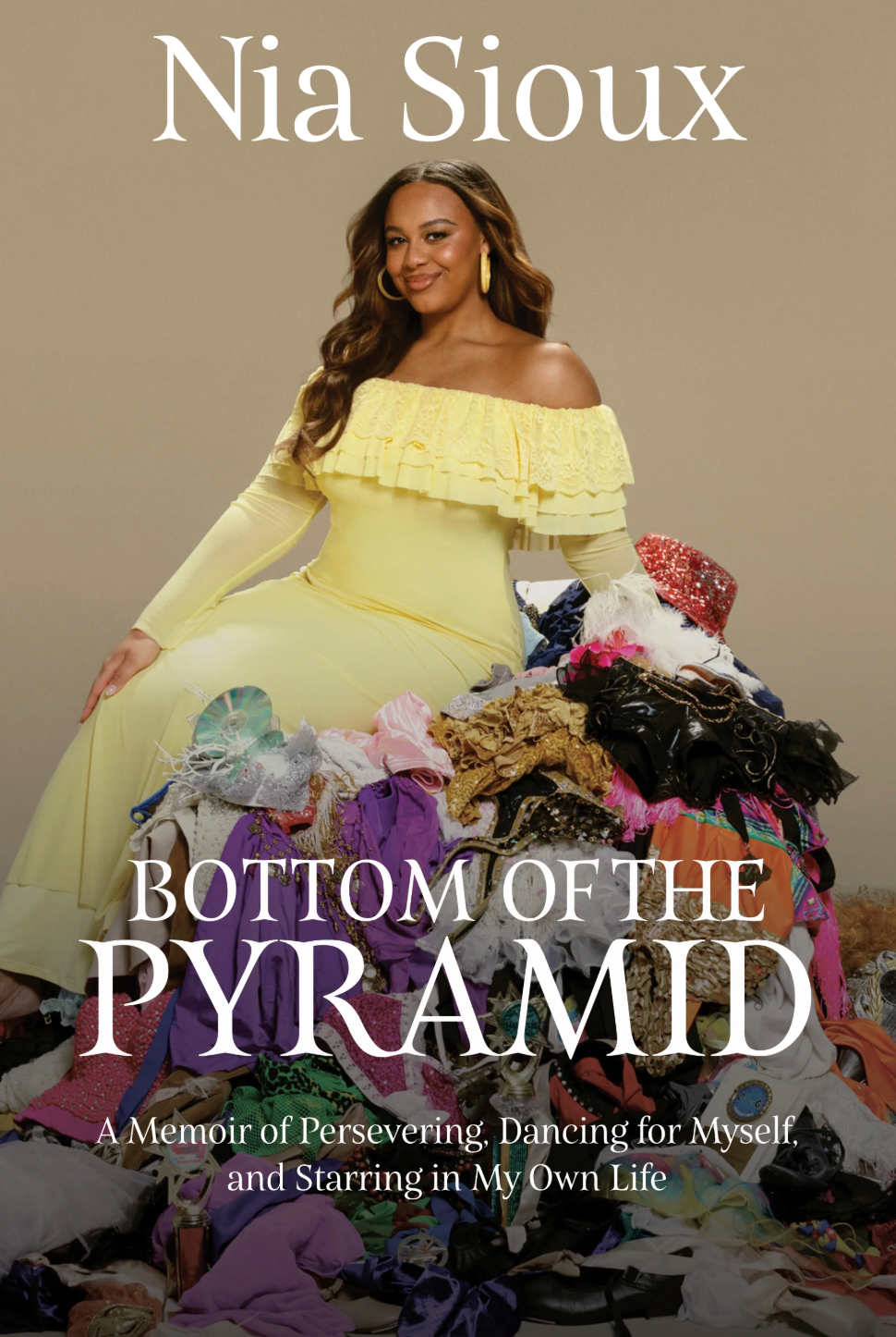 Book cover of Nia Sioux's memoir titled 'Bottom of the Pyramid,' featuring Nia Sioux in a yellow dress sitting beside a pile of colorful and patterned clothing and accessories.