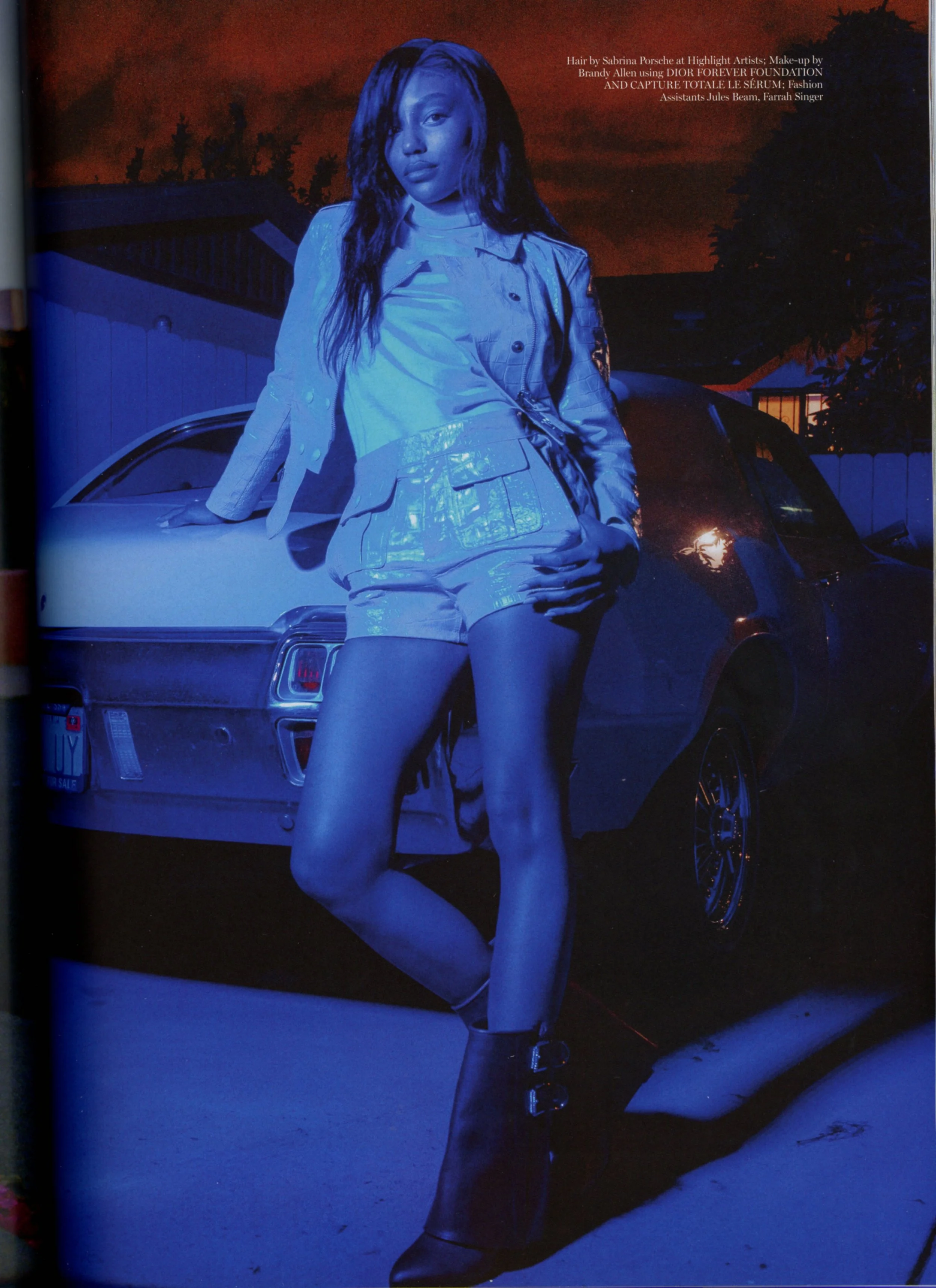 A woman with long curly hair standing outdoors near a car during a sunset, wearing a shiny jacket, shorts, and high-heeled boots.