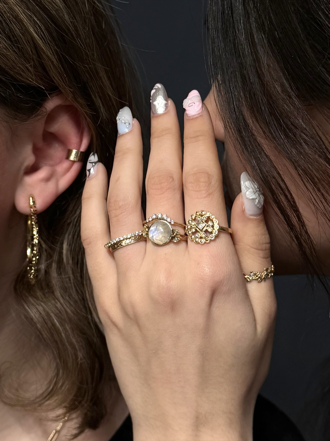 Close-up of a woman's hand with multiple rings, including a moonstone, gold and diamond rings, and gold jewelry, touching her face. She has floral nail art and wears gold earrings and a gold ear cuff.